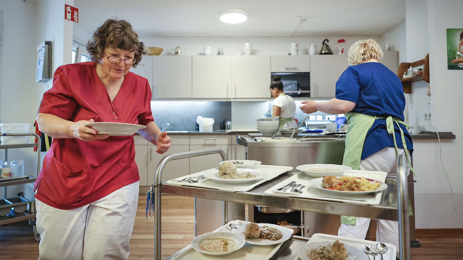 Jobcarving und Jobcoaching Zwei Pflegekräfte befüllen einen Servierwagen mit schön angerichteten Tellern mit Mittagessen.