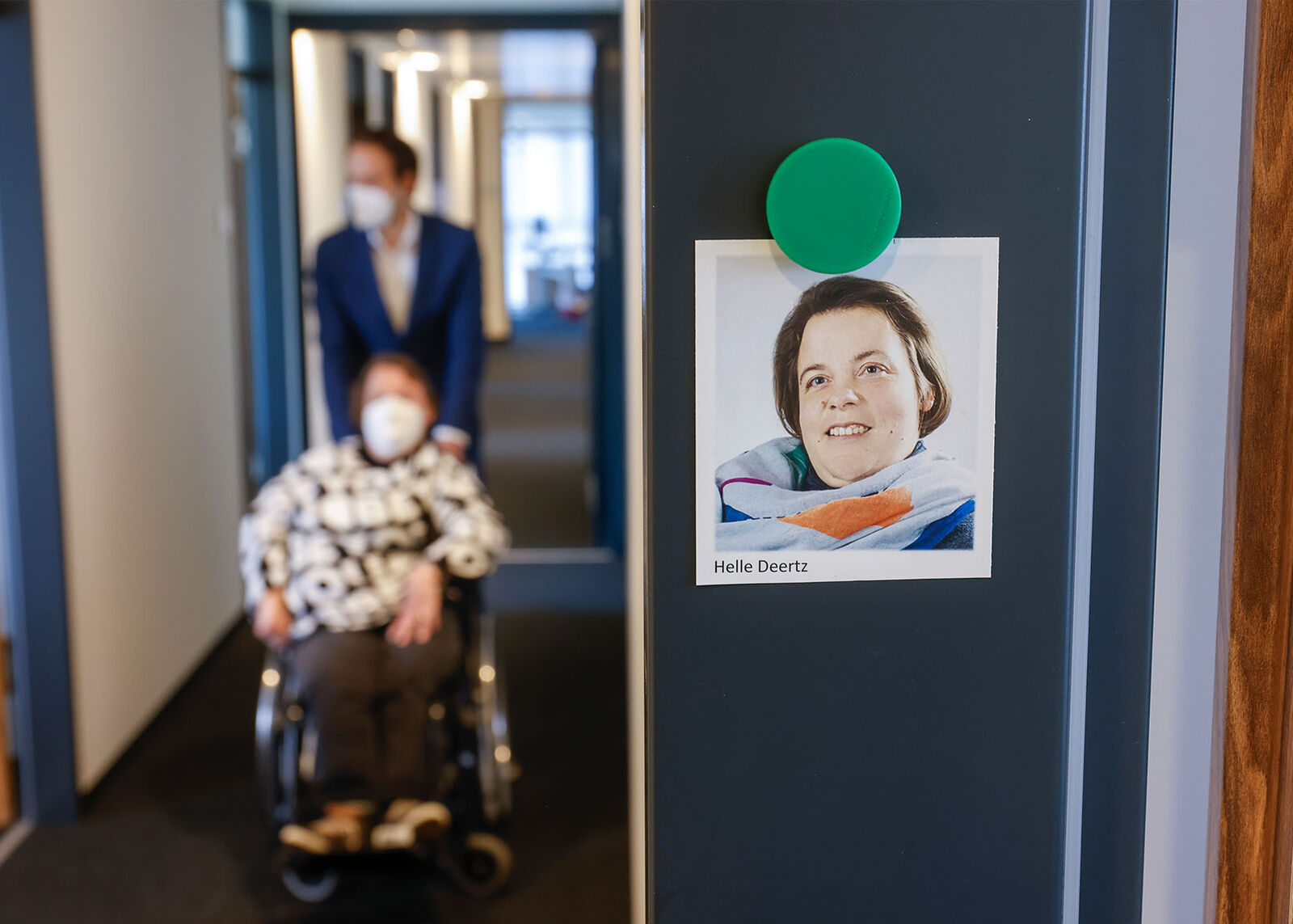 Arbeitsassistenz Helle Deertz wird von ihrem Assistenten im Rollstuhl über einen schmalen Gang geschoben. Am Türrahmen ihres Büros hängt ein Passbild von ihr.
