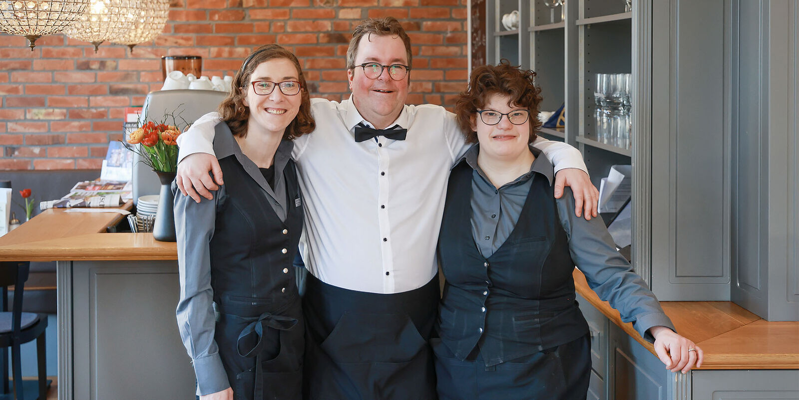 Hotel Villa Sterne Arne Krüger-Brörken mit zwei Kolleginnen Im Café steht Arne Krüger-Brörken Arm in Arm mit zwei Kolleginnen und lächelt in die Kamera.