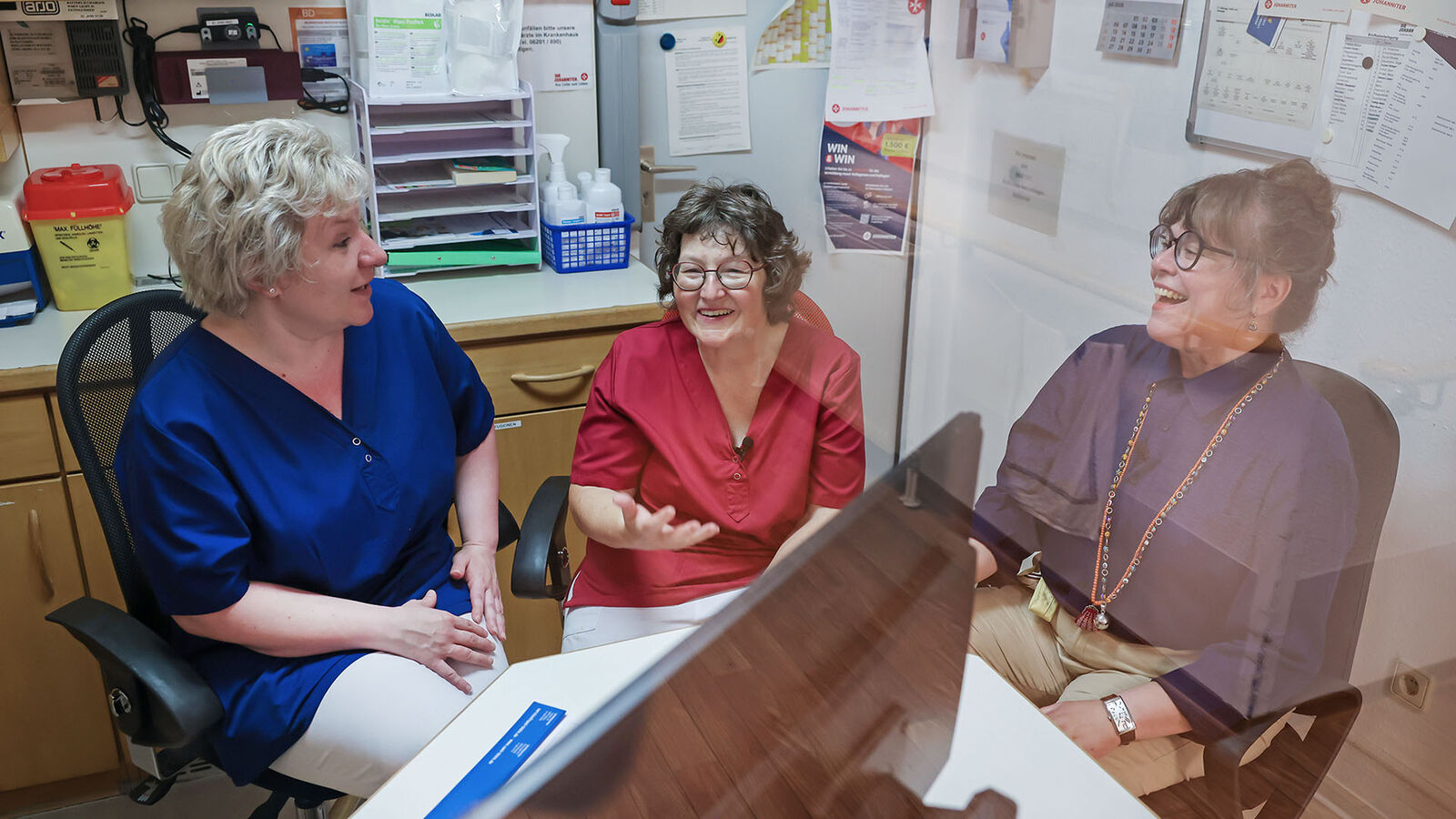 Jobcarving und Jobcoaching Drei Frauen mittleren Alters sitzen in einem Stationszimmer eines Pflegeheims und unterhalten sich lachelnd miteinander.