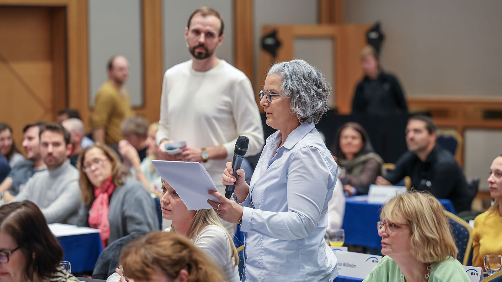 EAA-Netzwerktreffen, Fragen aus dem Publikum Eine Frau steht zwischen vielen Menschen, die an Tischen sitzen. Sie hält ein Manuskript in den Händen.