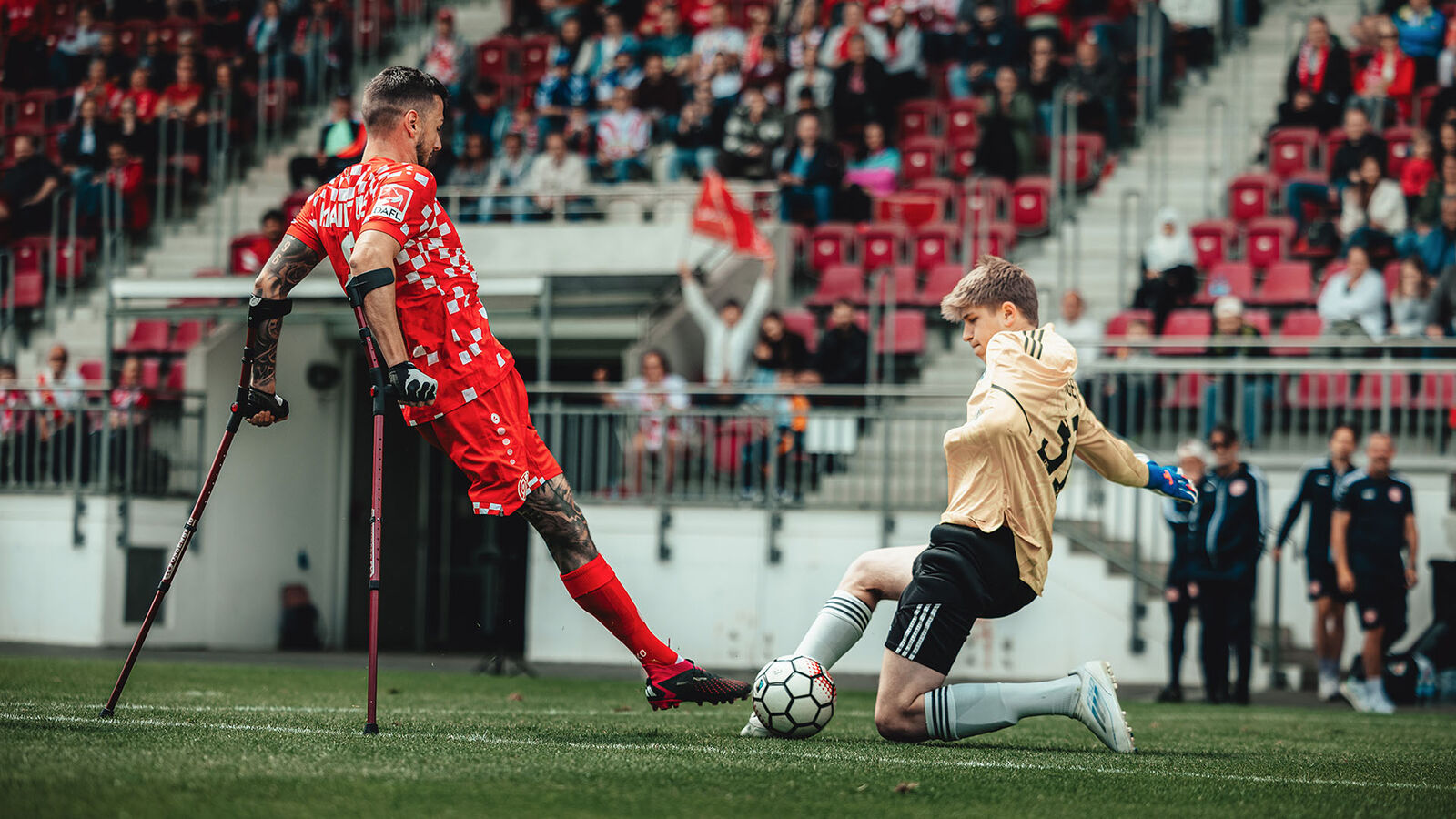 Ein Stürmer auf Krücken versucht noch mit de Fuß den Ball zu erreichen, während von der anderen Seite ein Torhüter mit nur einem Arm ebenfalls versucht den Ball mit dem Fuß wegzuspielen.