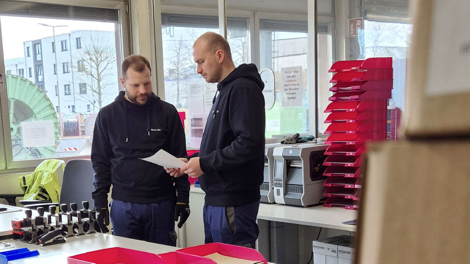 Zwei Männer stehen in einem Büro am Schreibtisch und unterhalten sich über ein Papier, das der rechte von ihnen in den Händen hält.