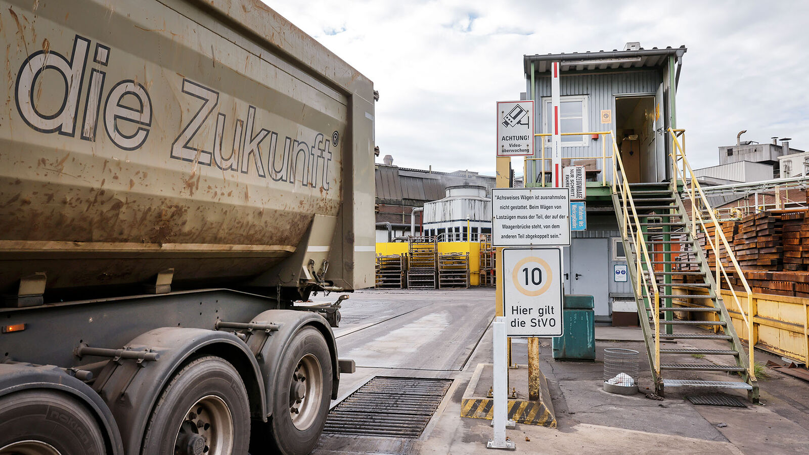 Eisenwerk Brühl, Lkw-Waage Ein Lkw mit der Aufschrift "Die Zukunft" setzt rückwärts auf die Waage des Eisenwerks Brühl. Rechts neben der Waage steht auf hohen Stahlträgern ein Baucontainer mit Büro. Zur geöffneten Tür führt eine lange Stahltreppe hinauf.