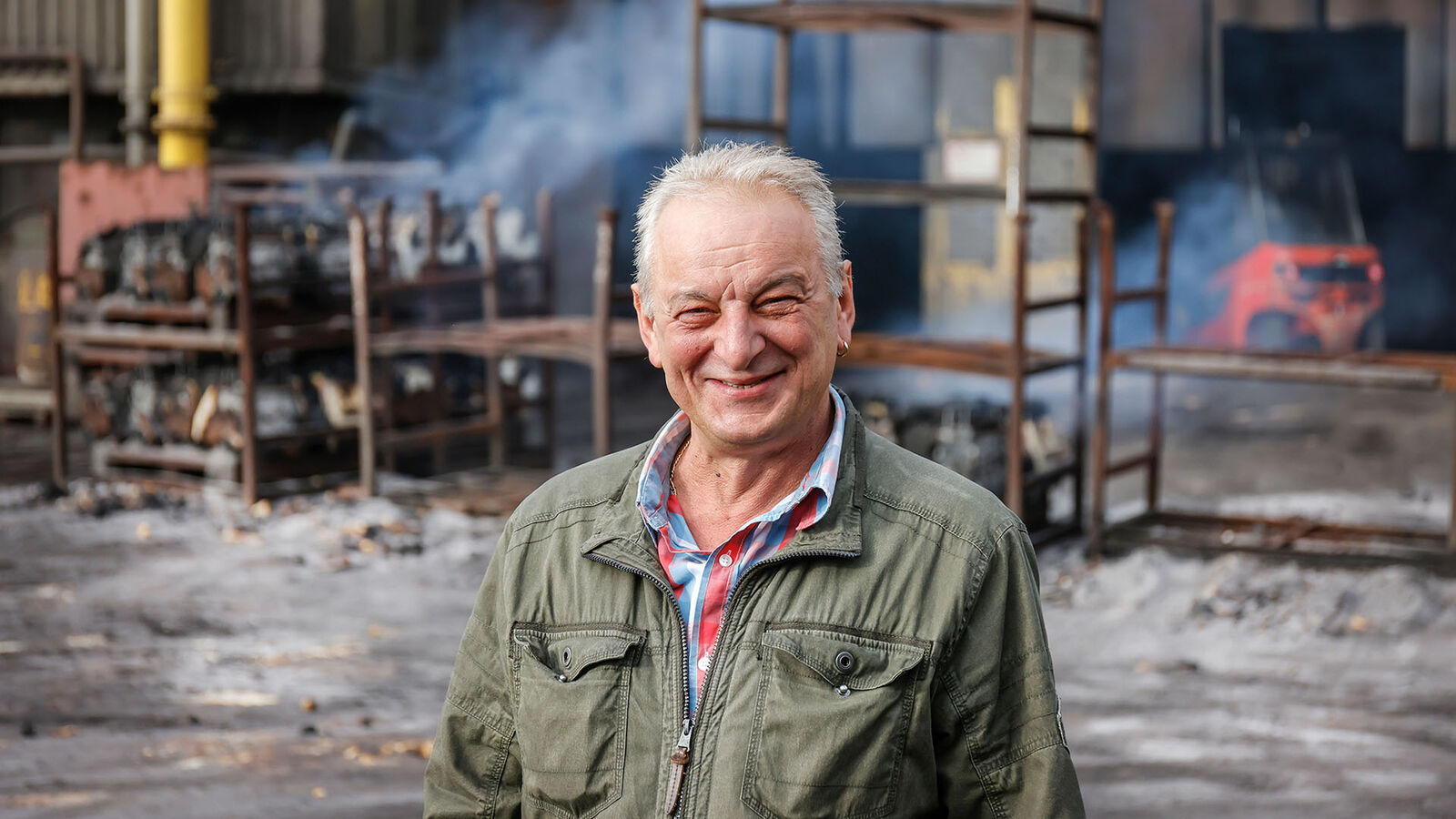 Eisenwerk Brühl, Frank Hofer Ein älterer Mann mit weißen Haaren und einigen Falten im Gesicht steht auf dem Hof des Eisenwerks vor verschieden hohen Stahlregalen.