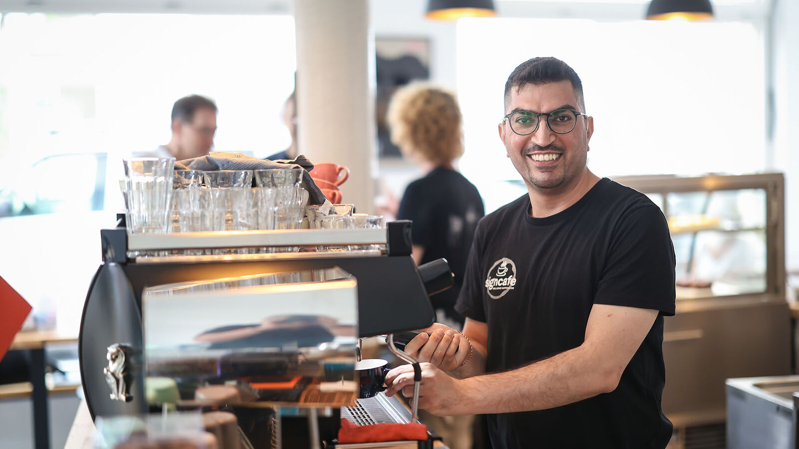 Ein junger Mann steht links von einer großen Siebträgerkaffeemaschine. Mit der rechten Hand Hand schiebt er eine blaue Tasse unter den Siebträger. 