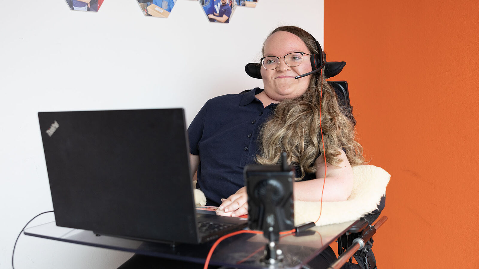 Patricia Lorenz sitzt in ihrem elektrischen Rollstuhl, vor ihr steht ihr Laptop auf einer am Rollstuhl befestigen Tischplatte und das Handy ist in einer Halterung vor ihr platziert.