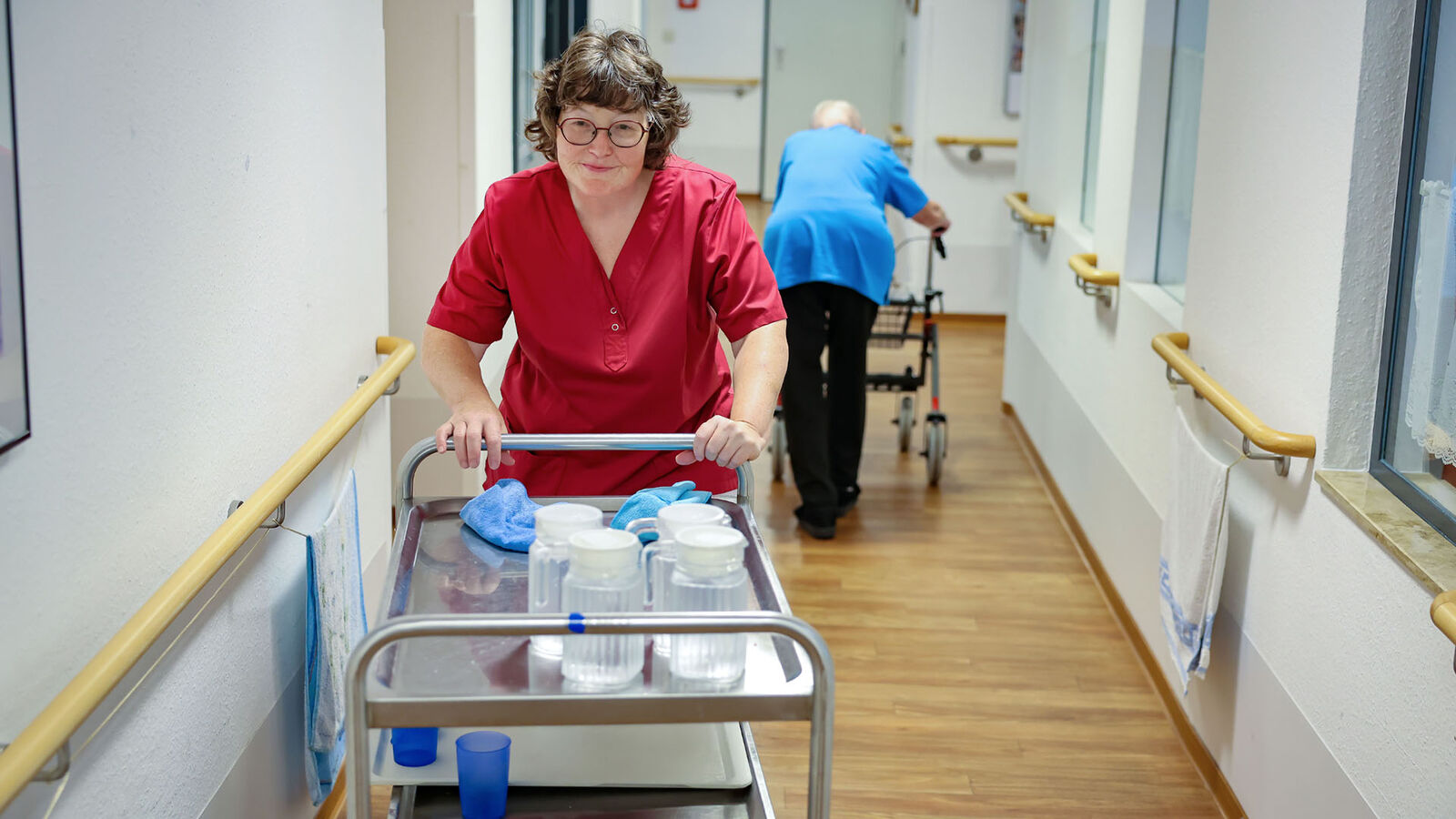 Jobcarving und Jobcoaching Eine weibliche Pflegekraft schiebt einen Servierwagen mit Wasserkaraffen über einen langen Gang.