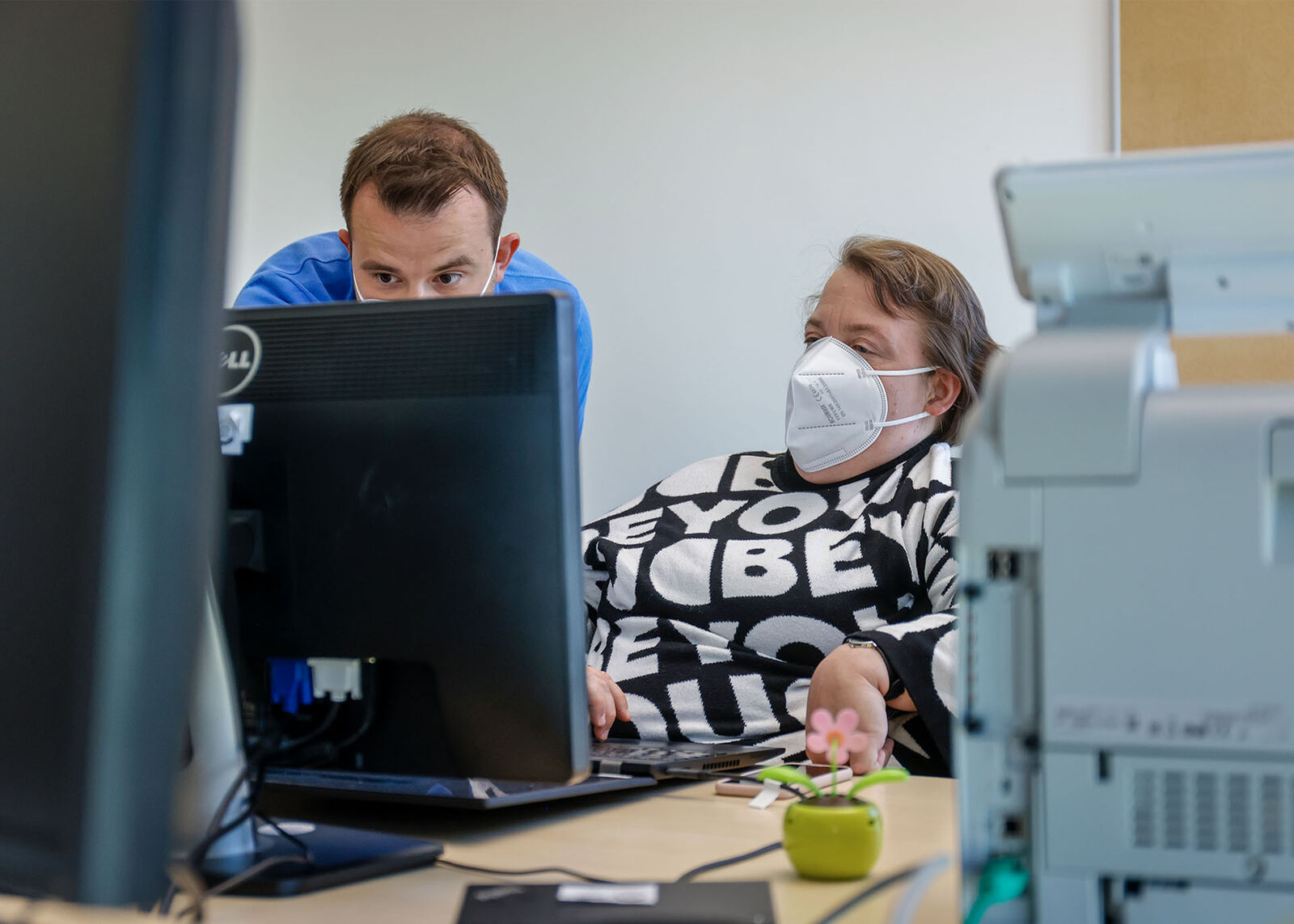 Arbeitsassistenz Eine Frau mit Mundschutz sitzt zurückgelehnt an ihrem PC. Ein Mann steht neben ihr und tippt auf der Tastatur.