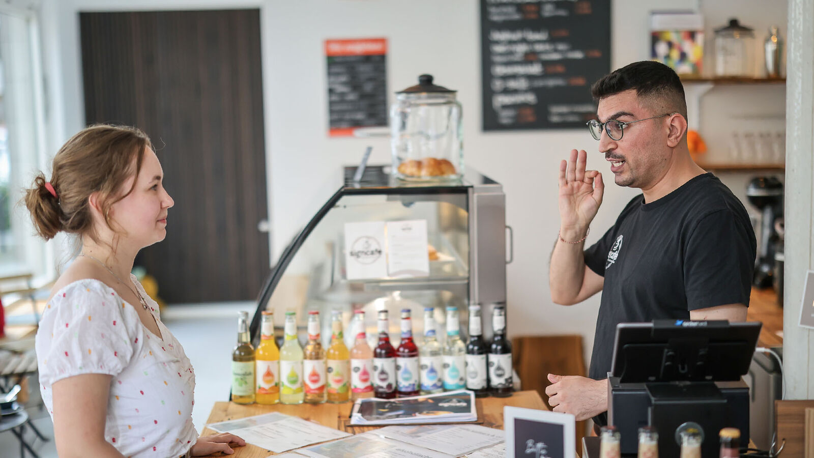In einem Café stehen sich eine junge Frau und ein junger Mann gegenüber. Die Frau versucht, in Gebärdensprache zu bestellen.