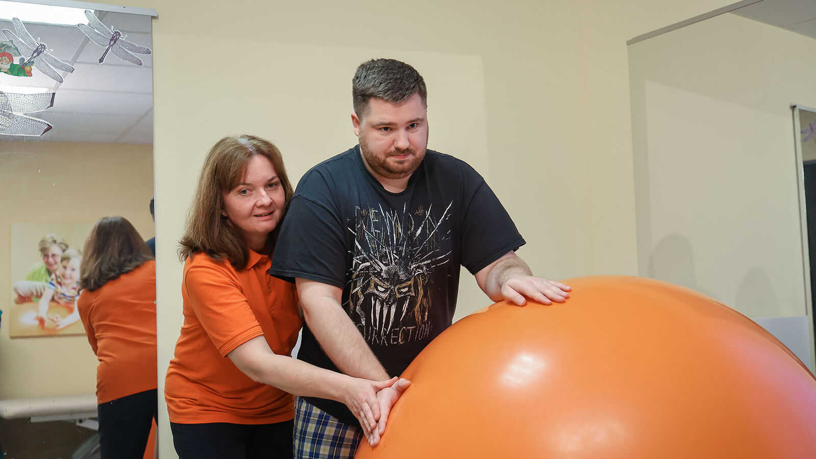 Physiotherapie mit großem Gymnastikball Eine Physiotherapeutin behandelt einen jungen Mann, der sich auf einen großen Gymnastikball abstützt. Die Therapeutin steht hinter ihm.