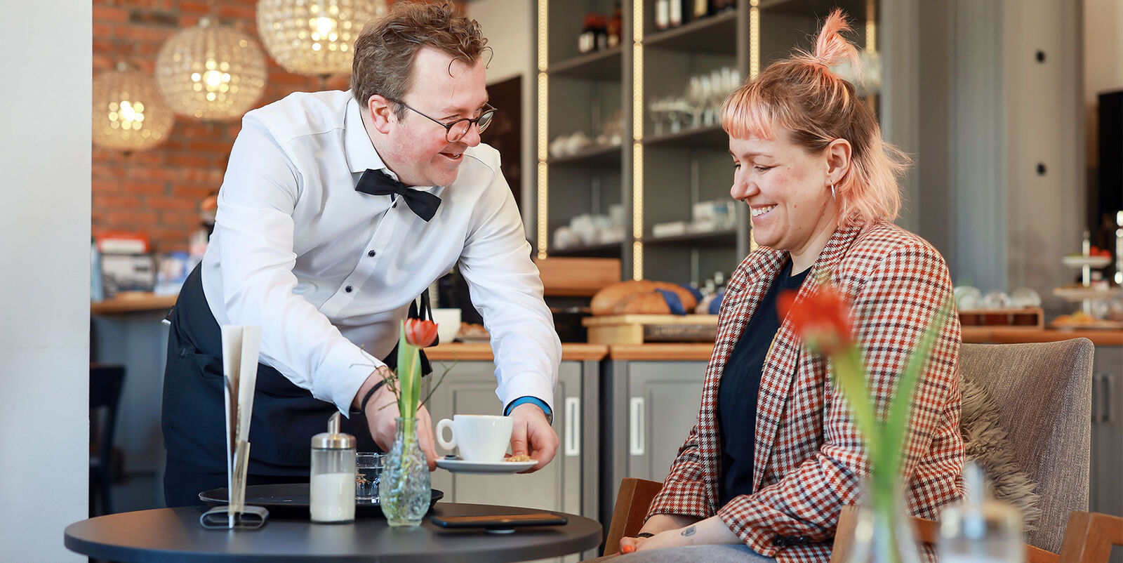 Arne Krüger-Brörken mit Gast Arne Krüger-Brörken stellt eine Kaffeetasse auf einen Cafétisch vor einem Gast ab.