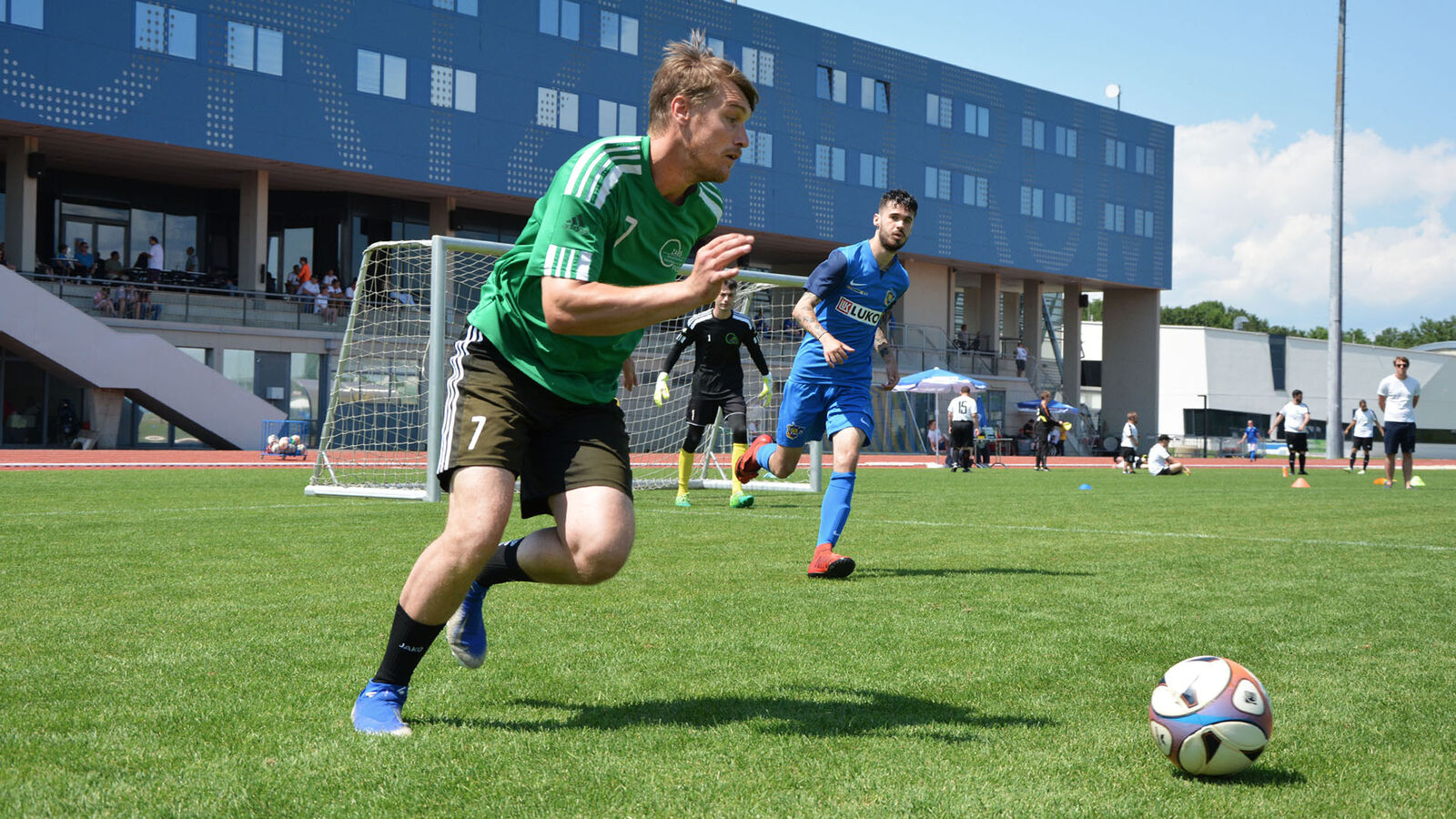 Vor einem Tor rennt ein Fußballer in grünschwarzem Trikot rennt auf einen Ball zu. Der Torwart steht bereit und ein Verteidiger läuft auf den Ball zu.