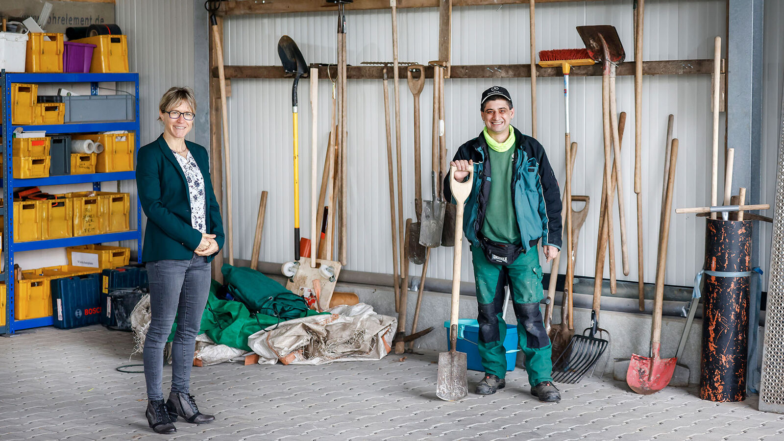 GaLa Service Inklusionsbetrieb Eine schlanke Frau in Jeans und Blazer steht neben einem Mann mittleren Alters in Arbeitskleidung in einer Halle. An den Wänden hängen Gartenwerkzeuge.