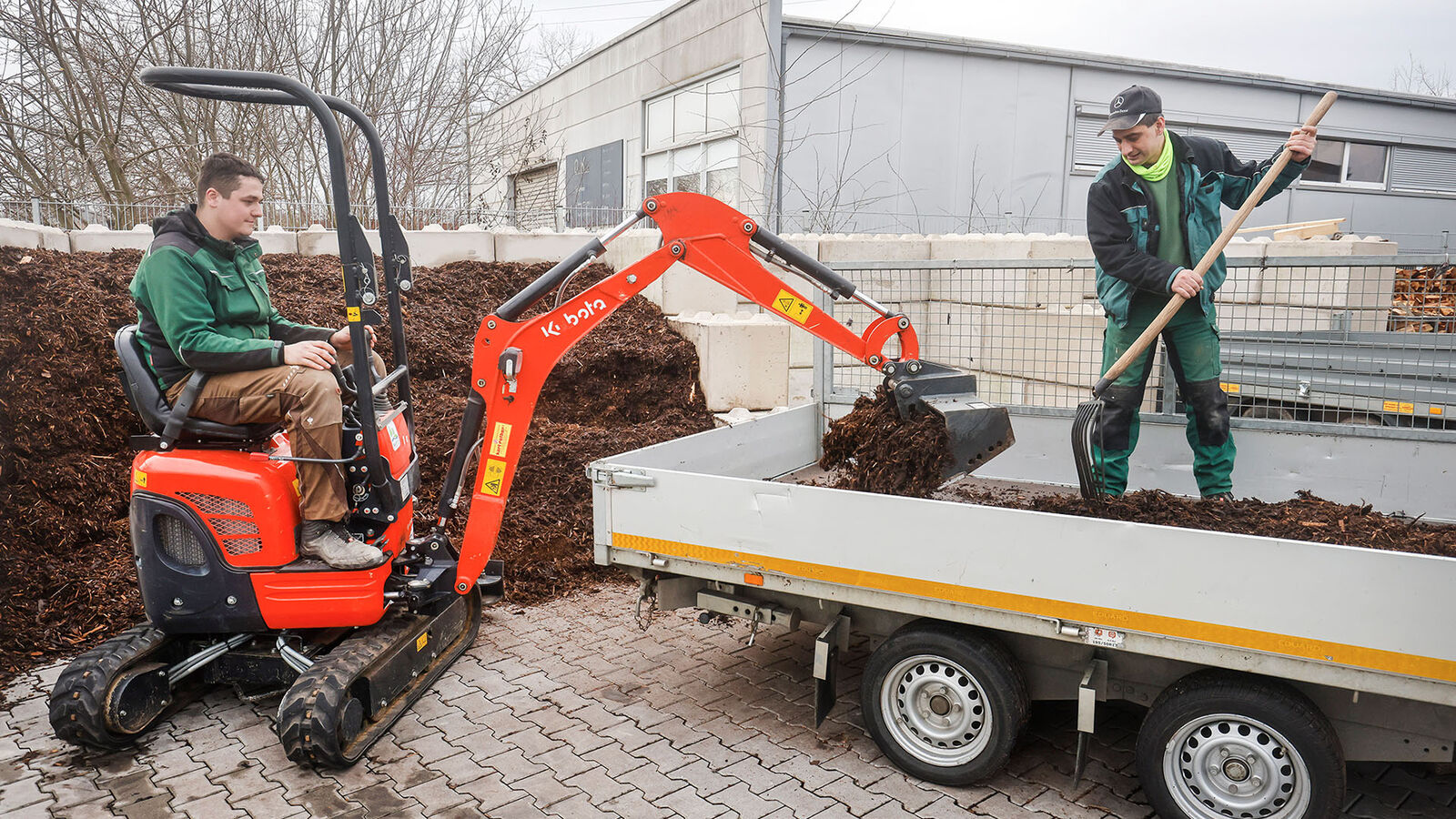 GaLa Service Inklusionsbetrieb Ein kleiner Radlader mit Schaufel steht an einem offenen Pritschenwagen und lädt Erde auf. Auf der Pritsche steht ein Gartenarbeiter mit einer Schaufel und verteilt die Erde.