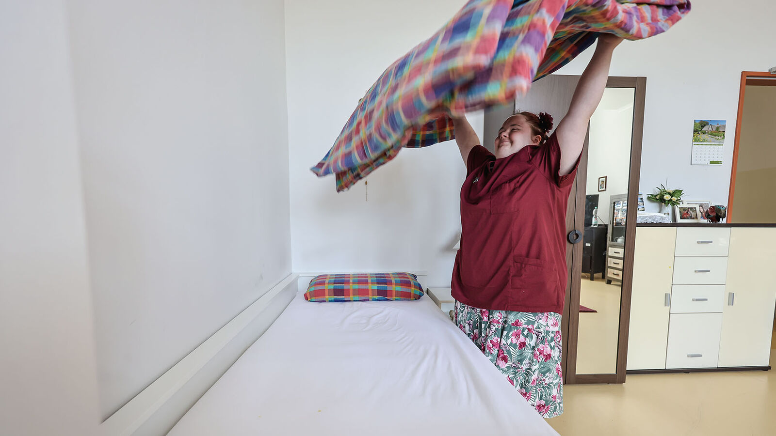 Franziska Wolf beim Bettenmachen. Die kleine rundliche Frau schüttelt das karierte Oberbett hoch über ihrem Kopf aus.