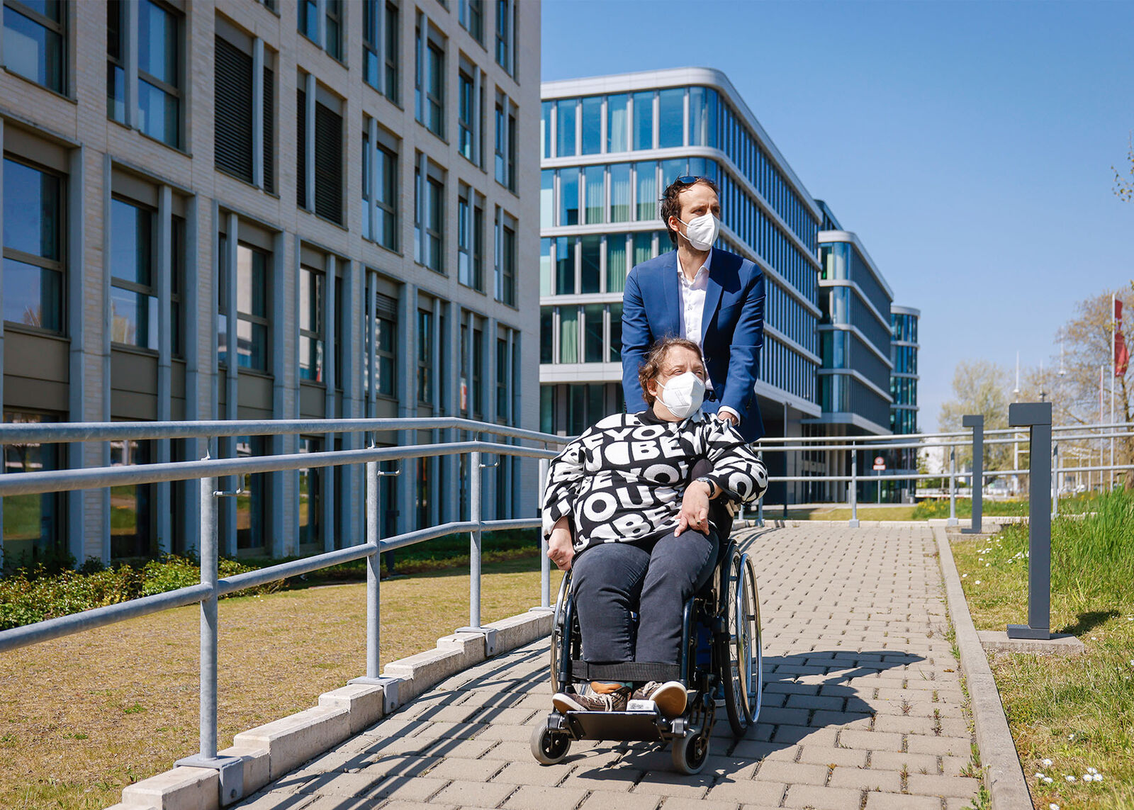 Arbeitsassistenz Helle Deertz wird von ihrem Assistenten im Rollstuhl vor dem Gebäude der GIZ über einen gepflasterten Weg geschoben.
