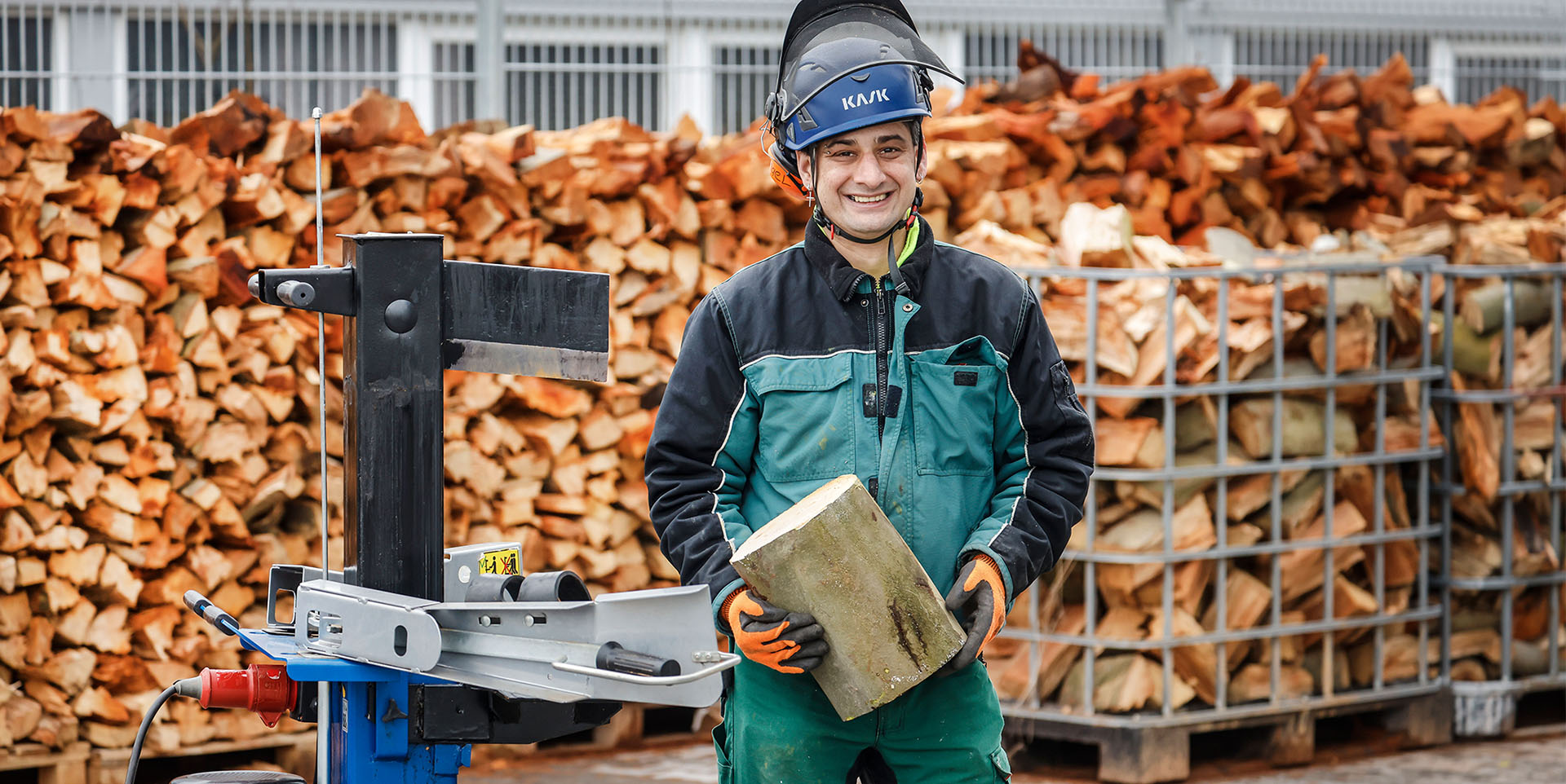 GaLa Service Inklusionsbetrieb Ein Mann mittleren Alters im Arbeitsoverall und Schutzhelm mit aufgeklapptem Visier steht mit einem Holzblock vor einem großen Brennholzstapel.