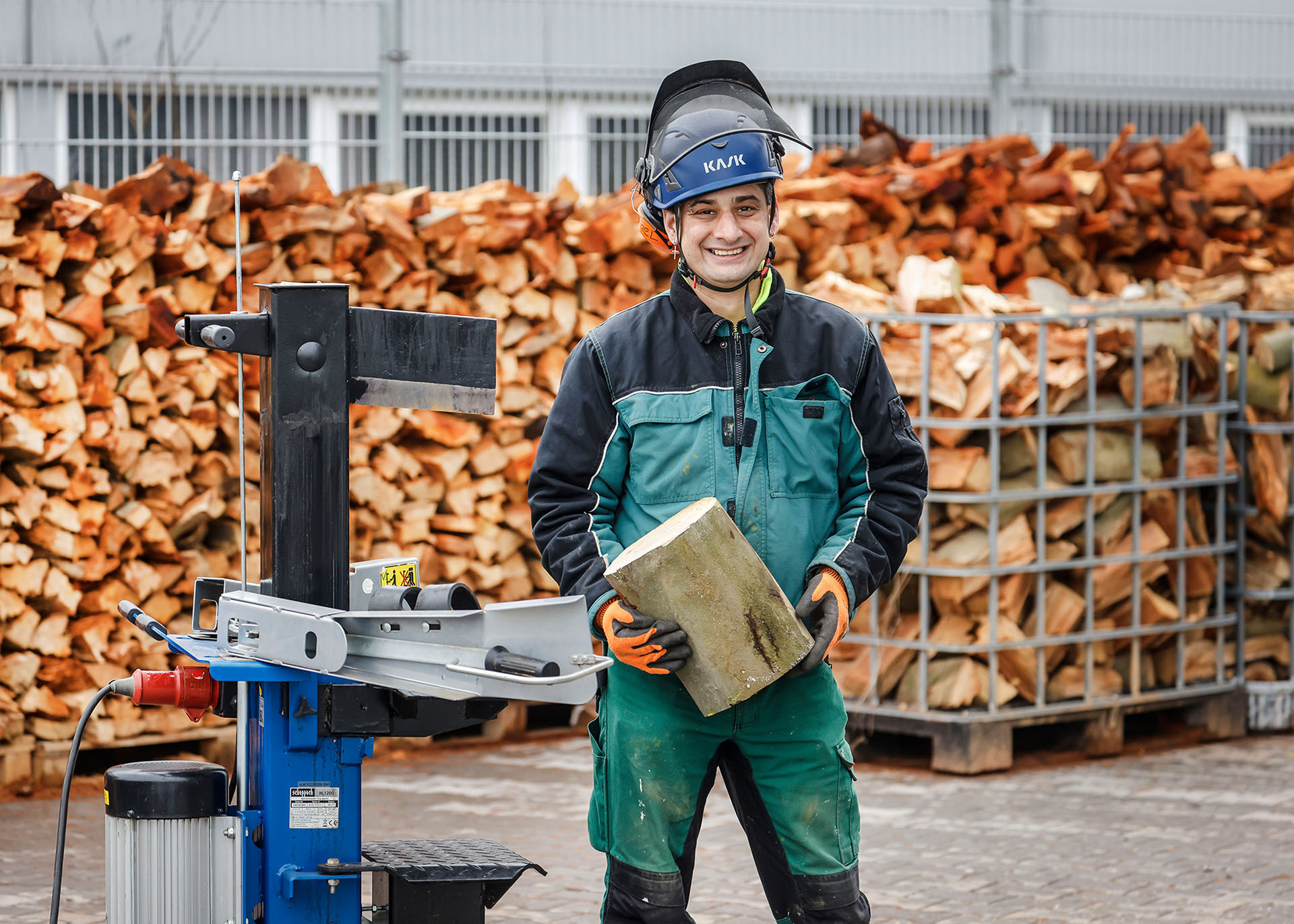 Ein Mann mittleren Alters im Arbeitsoverall und Schutzhelm mit aufgeklapptem Visier steht mit einem Holzblock vor einem großen Brennholzstapel.