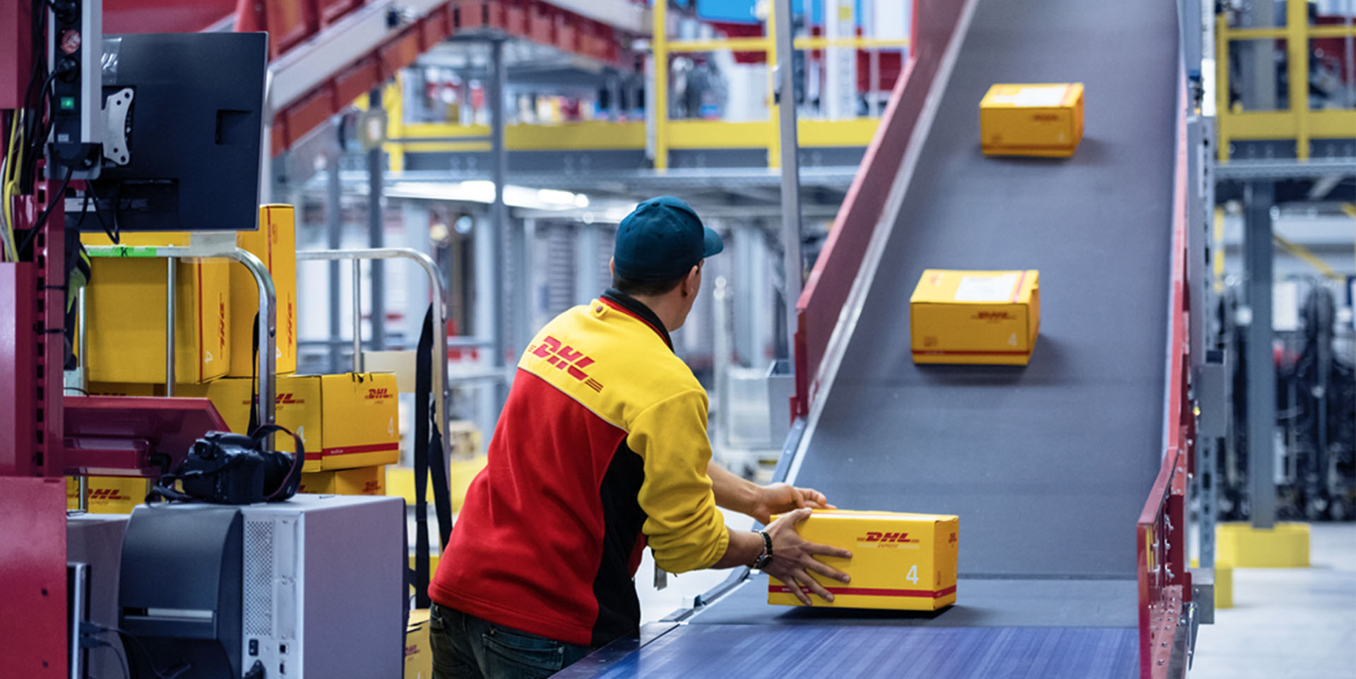Ein Mitarbeiter in gelbroter Jacke mit der Aufschrift „DHL“ legt Paket auf ein Fließband.