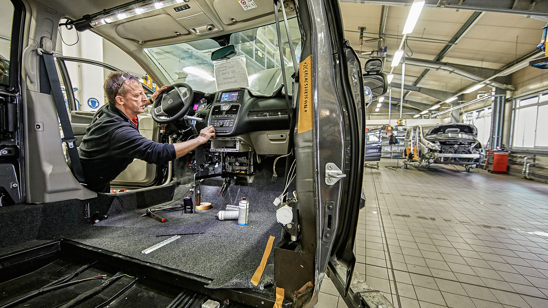 Ein Hochdachkombi steht mit geöffneten Türen in einer großen Halle. Durch die Fahrertür lehnt sich ein älterer Monteur in den Wagen und befestigt etwas rechts neben dem Lenkrad. Das Auto ist innen komplett leer. Der Blick fällt durch die Schiebetür auf der Beifahrerseite.