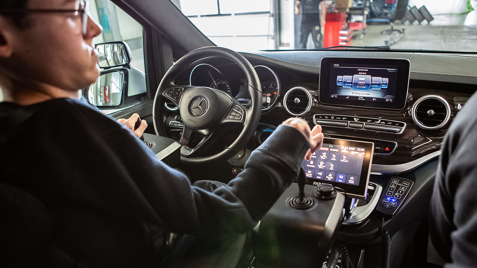 Ein Mann mittleren Alters mit deformierten Händen sitzt am Lenkrad eines modernen Pkws. Unterhalb des Fahrzeug-Displays sind ein zusätzliches Tablett und zwei Joysticks angebracht. Damit steuert der Mann das Auto.