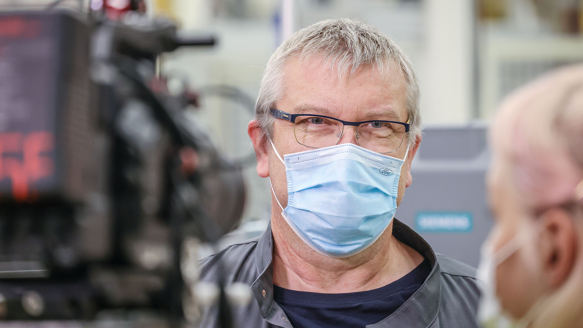 Im Vordergrund des Bildes ist eine Videokamera im Anschnitt zu sehen. Sie zeigt auf einen Mann zwischen 50 und 60 Jahren. Er wird von einer jungen Frau interviewt. Beide tragen Coronamasken.