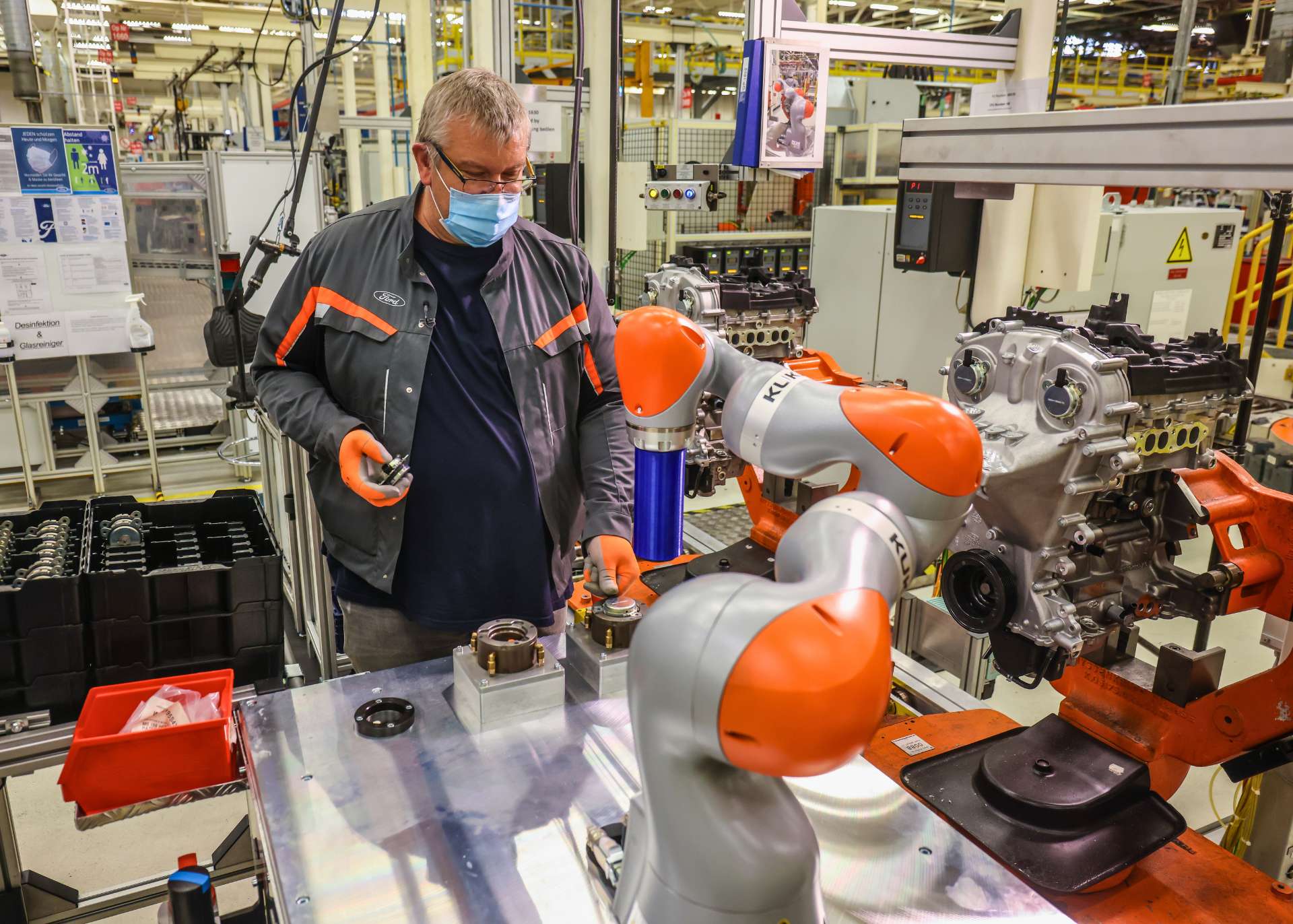 Der Blick fällt von vorn auf einen Arbeitsplatz, an dem ein Mensch und ein Roboter gemeinsam arbeiten. Der Roboter ist auf einem Tisch montiert und so hoch wie der Oberkörper des Mannes.