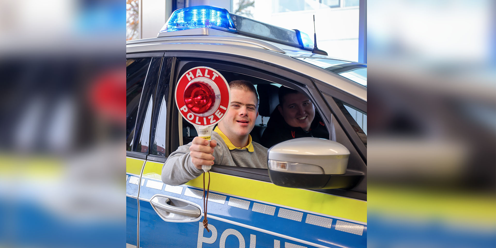 Ein junger Mann mit Downsyndrom sitzt im Beifahrersitz eines Polizeifahrzeugs. Er hält eine rote Kelle aus dem geöffneten Fenster und lacht.