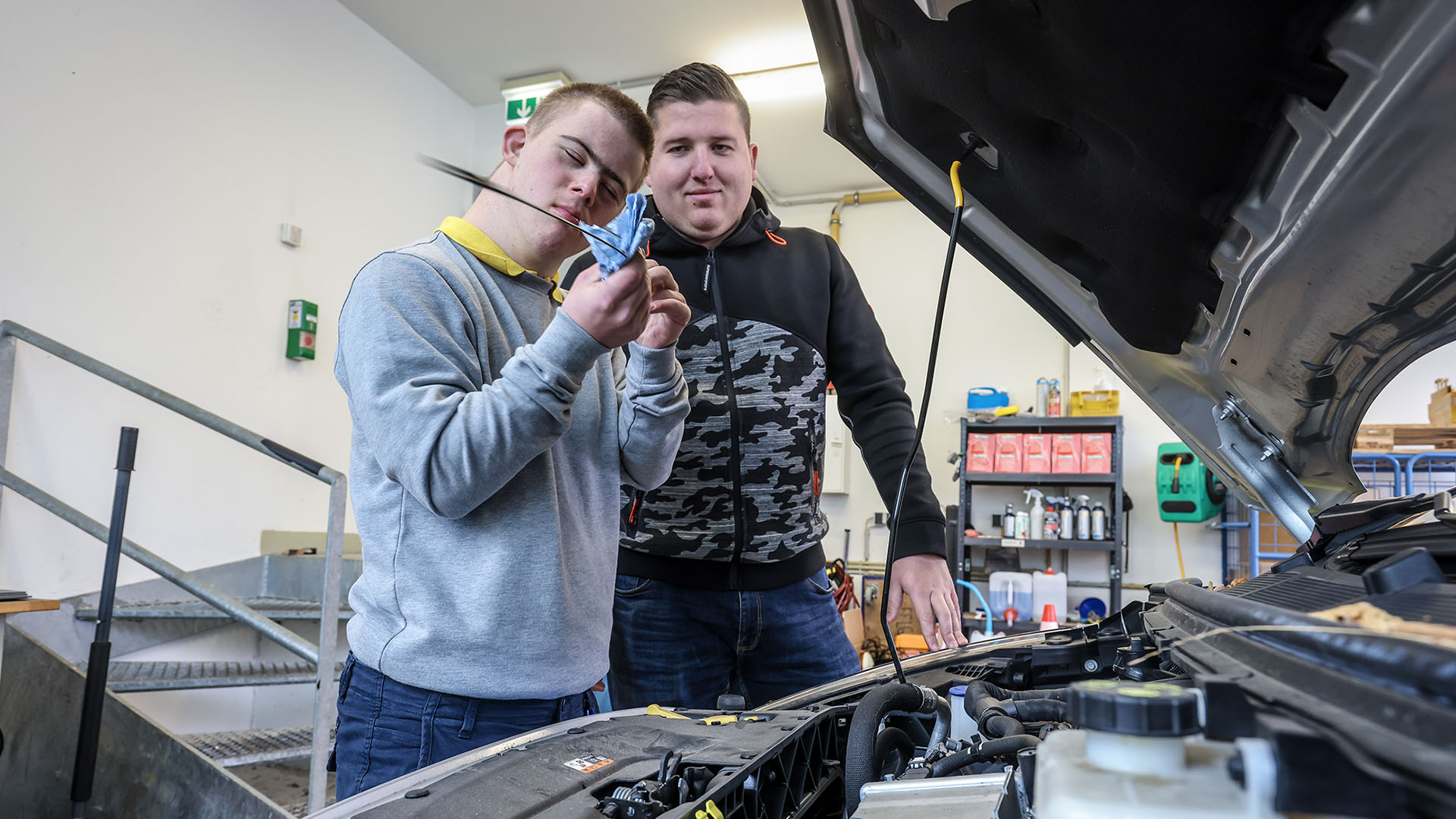 Ein junger Mann mit Downsyndrom prüft den Ölstand eines Kfz. Ein Mann mittleren Alters schaut ihm dabei über die Schulter.