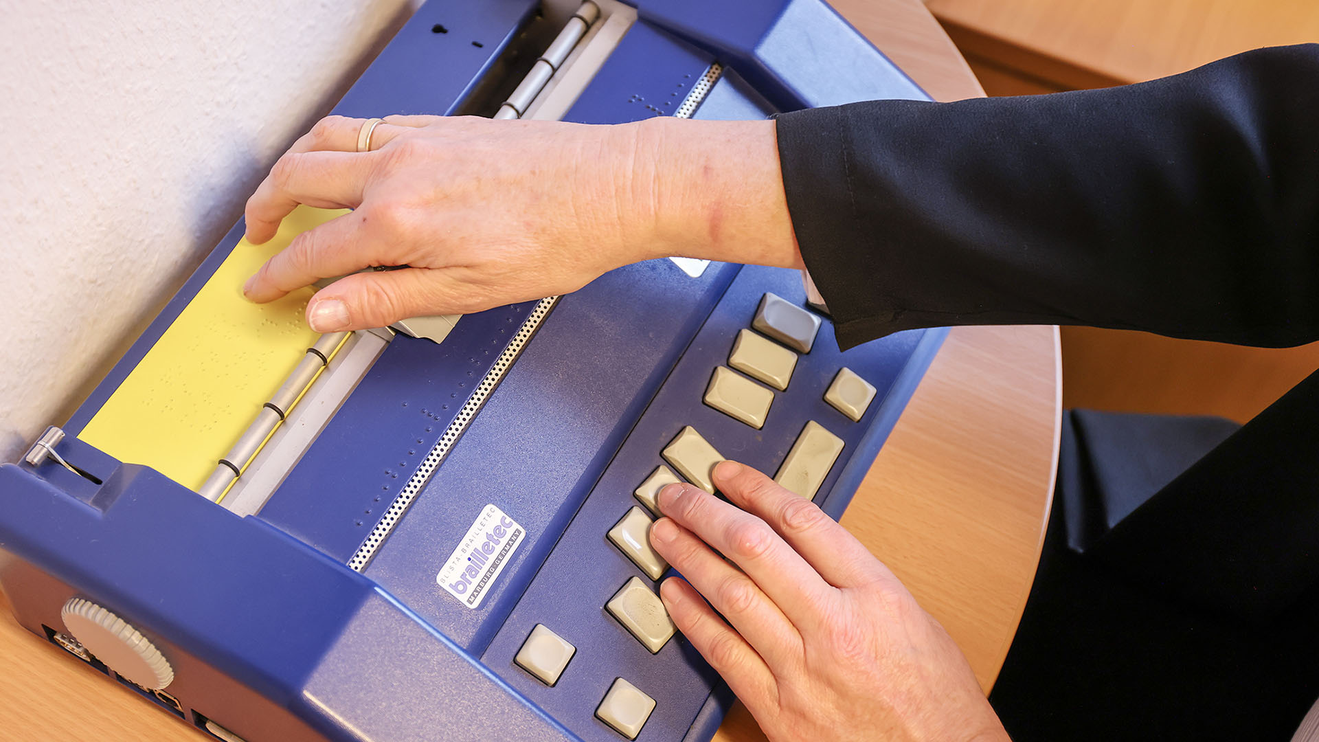 Aus einem blauen flachen Drucker mit ergonomisch angeordneten großen Tasten ragt ein gelbes Papier heraus. Es ist mit reliefartiger Brailleschrift bedruckt.