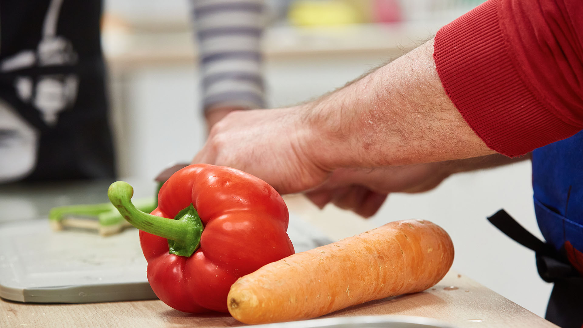Eine große Karotte und eine pralle rote Paprika liegen auf einer Arbeitsplatte. Von rechts greift eine Männerhand ins Bild.