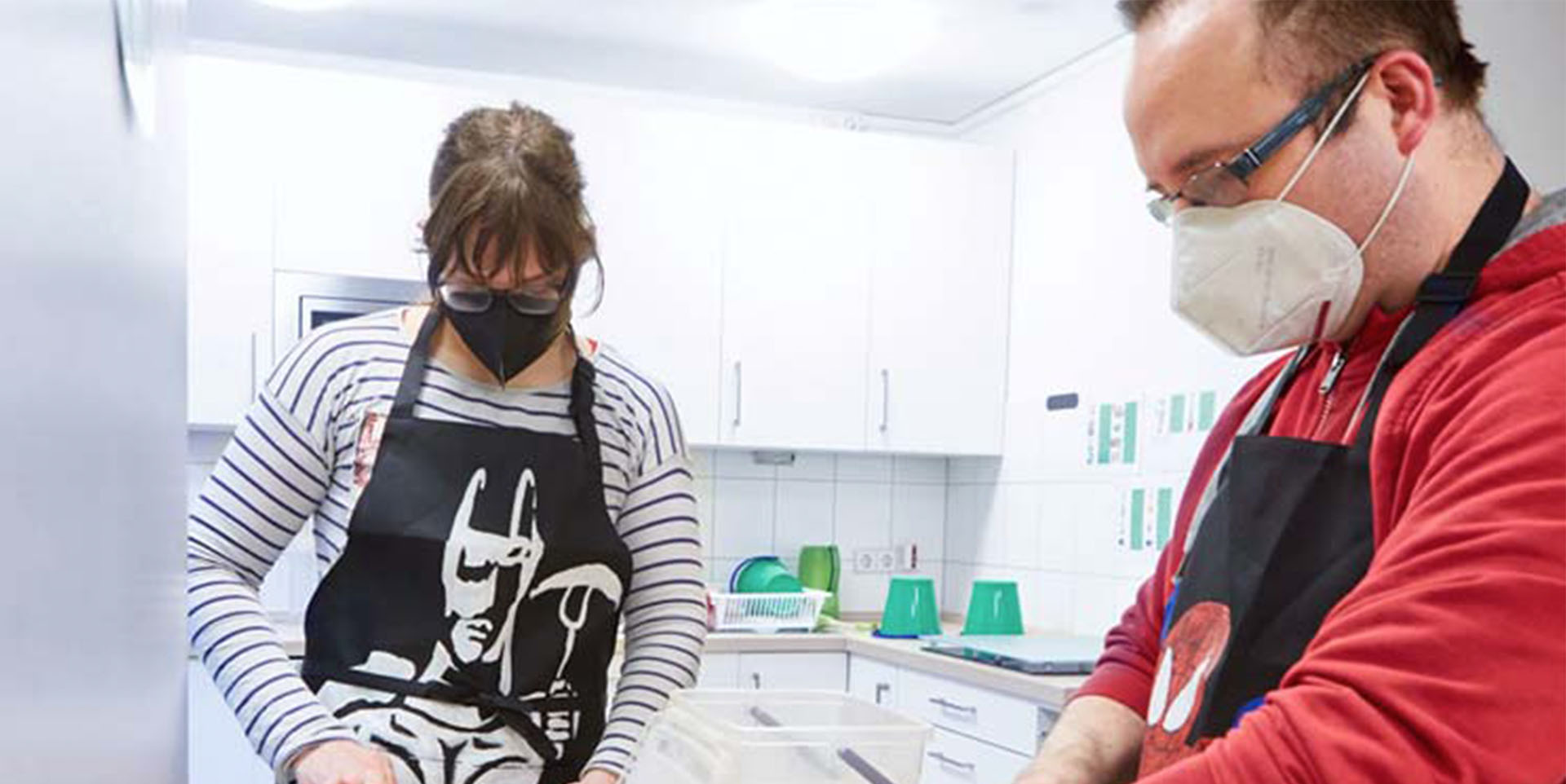 Eine junge Frau mit schwarzer Schürze und Gesichtsmaske sowie ein Mann mittleren Alters im roten Pulli und schwarzer Schürze bei der Küchenarbeit.