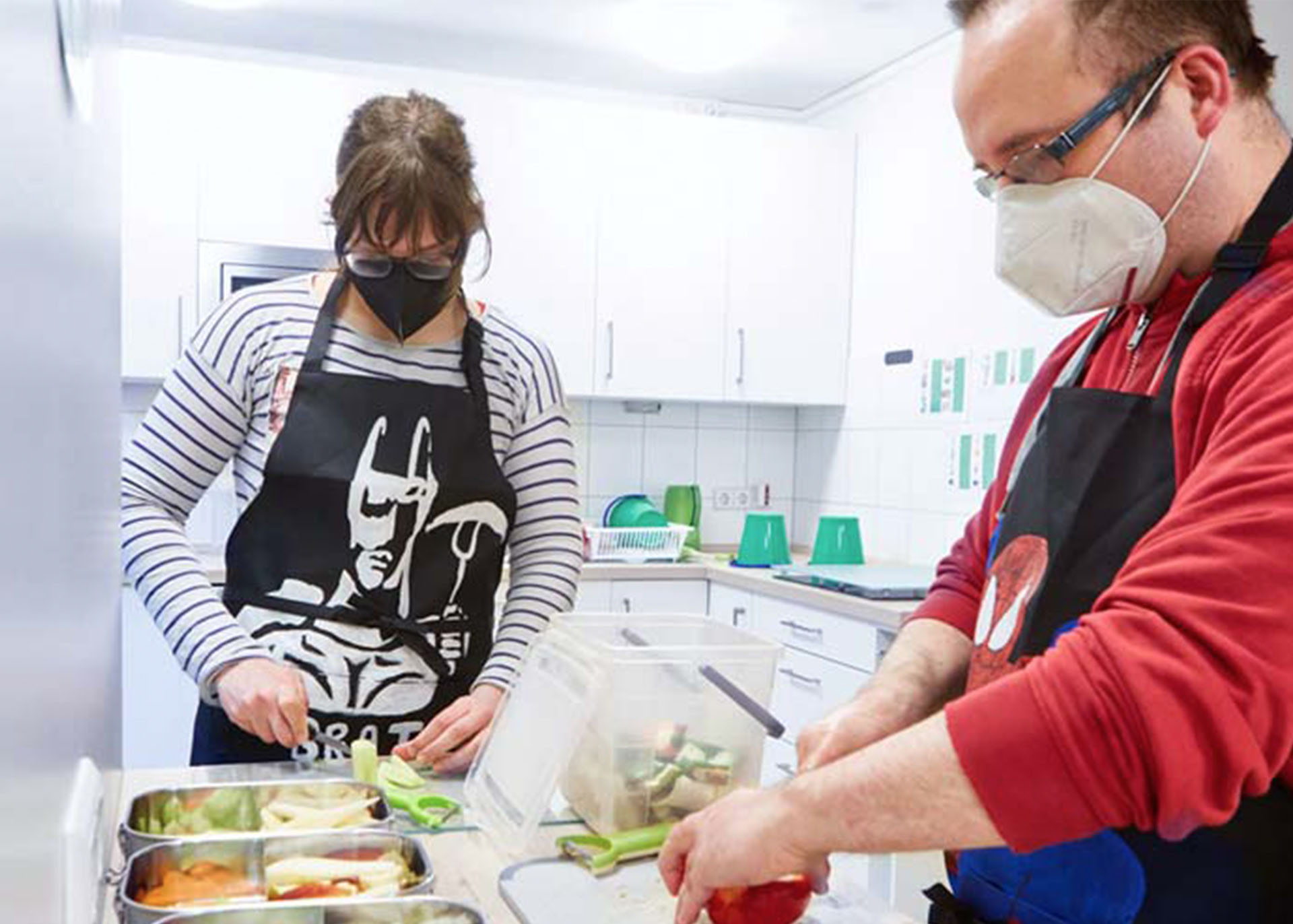 Eine junge Frau mit schwarzer Schürze und Gesichtsmaske sowie ein Mann mittleren Alters im roten Pulli und schwarzer Schürze bei der Küchenarbeit.
