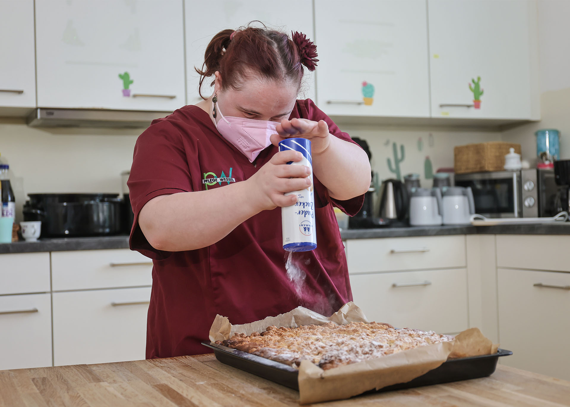 Franziska Wolf streut aus einer großen runden Dose Puderzucker auf ein Bleck mit frisch gebackenem Obststreuselkuchen.