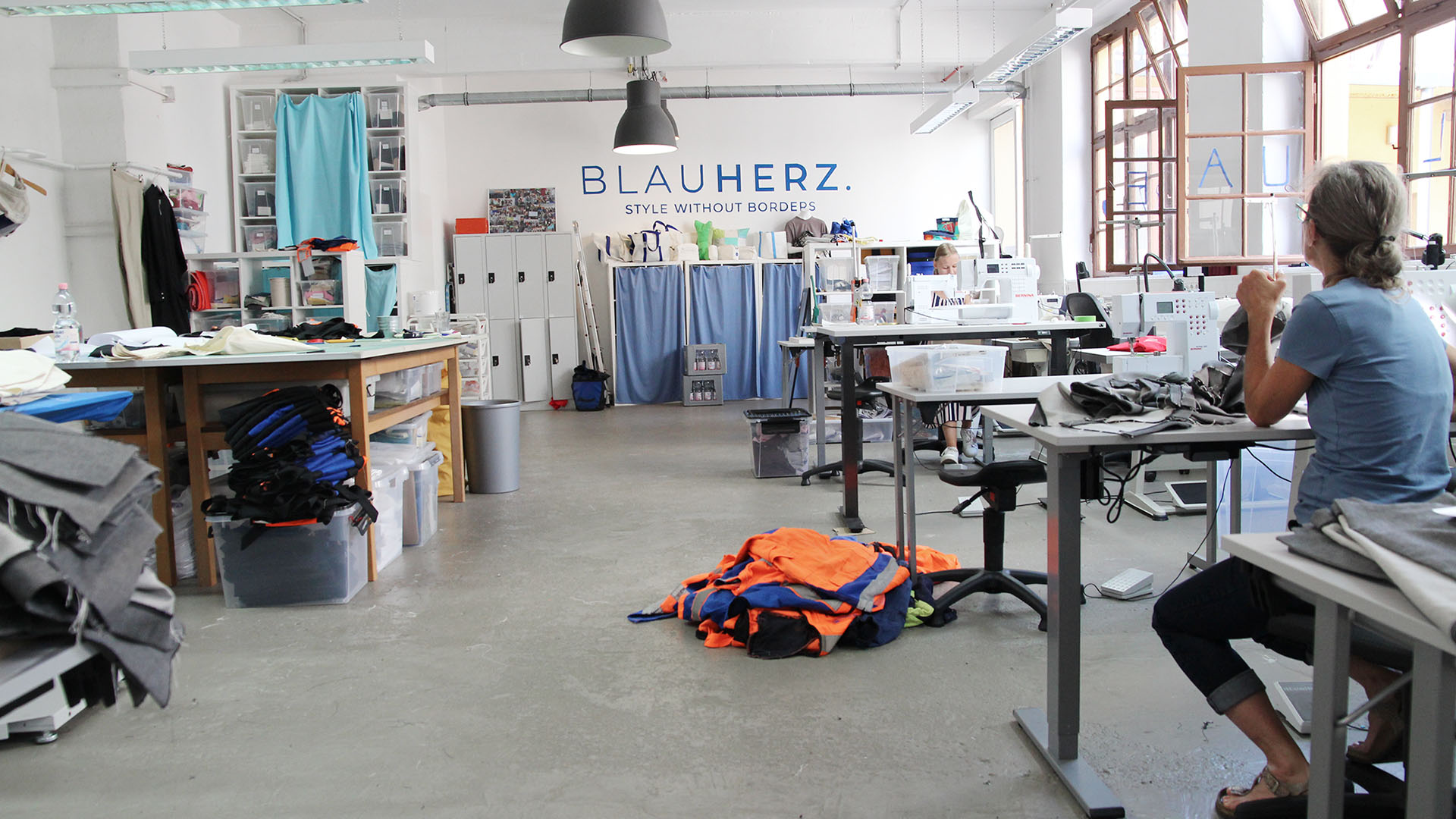Das Blauherz-Atelier mit dem großen Zuschneidetisch links und vier Näharbeitsplätzen rechts. Vor Kopf ist das Regal mit der aktuellen Taschenkollektion.
