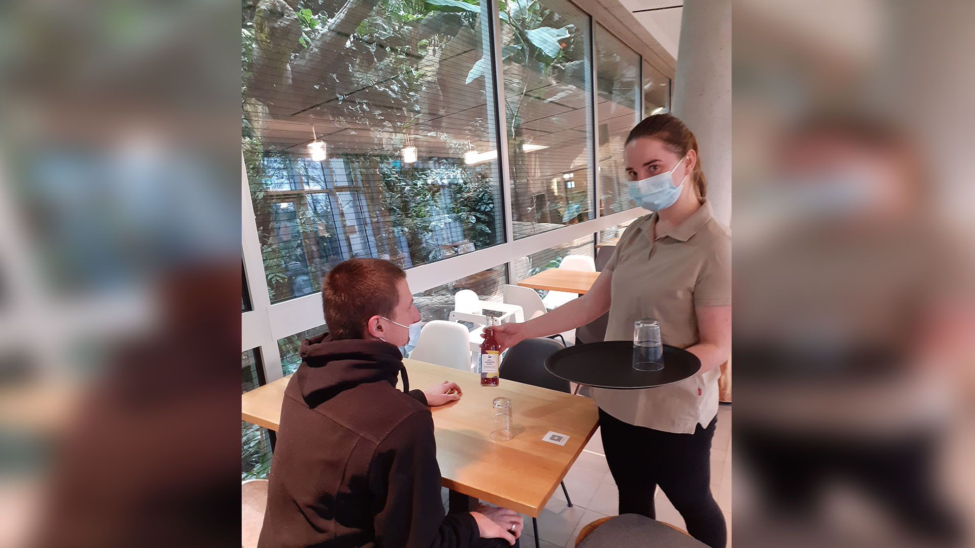 Eine junge Frau mit Mundschutz kellnert in einem hell möblierten Restaurant. Durch die hohen Scheiben sind tropische Grünpflanzen zu sehen.