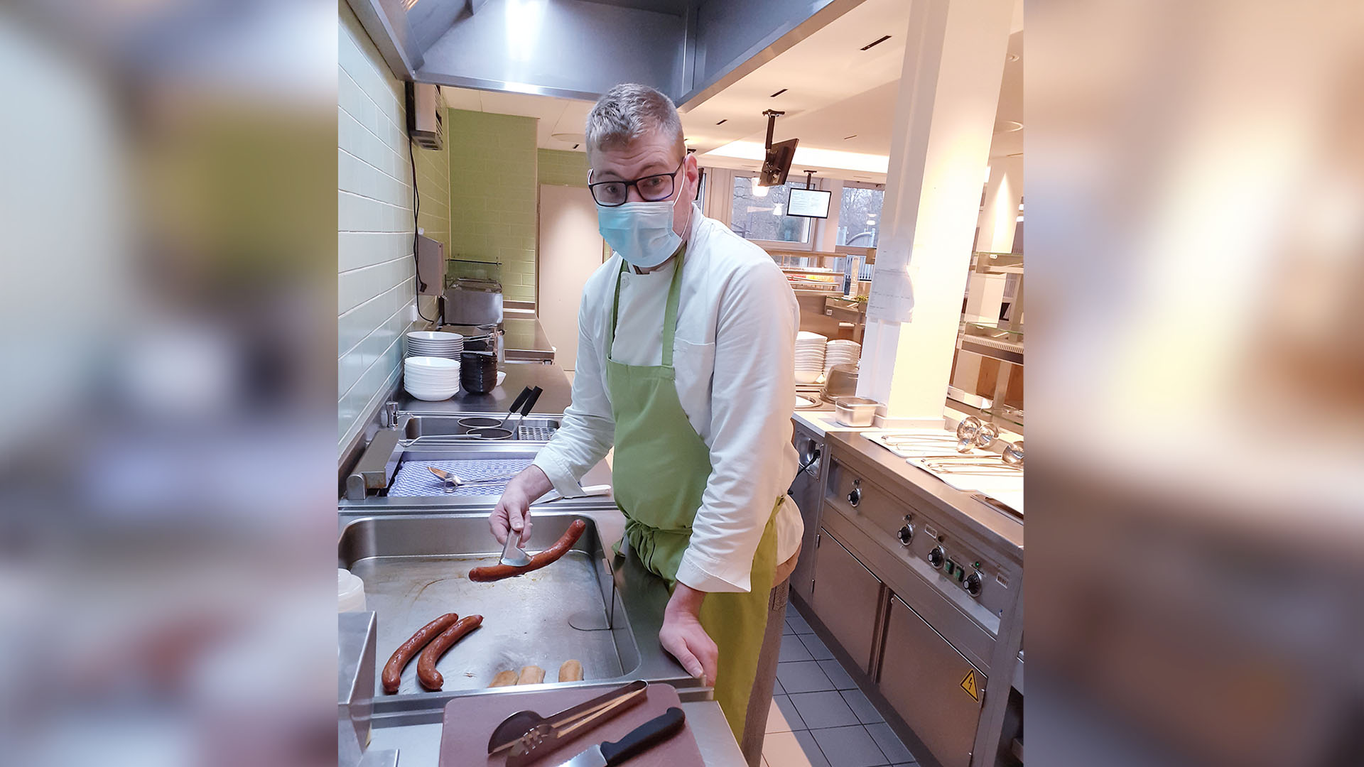 Ein Mann mittleren Alters steht in einer Gastroküche und wendet Würstchen.