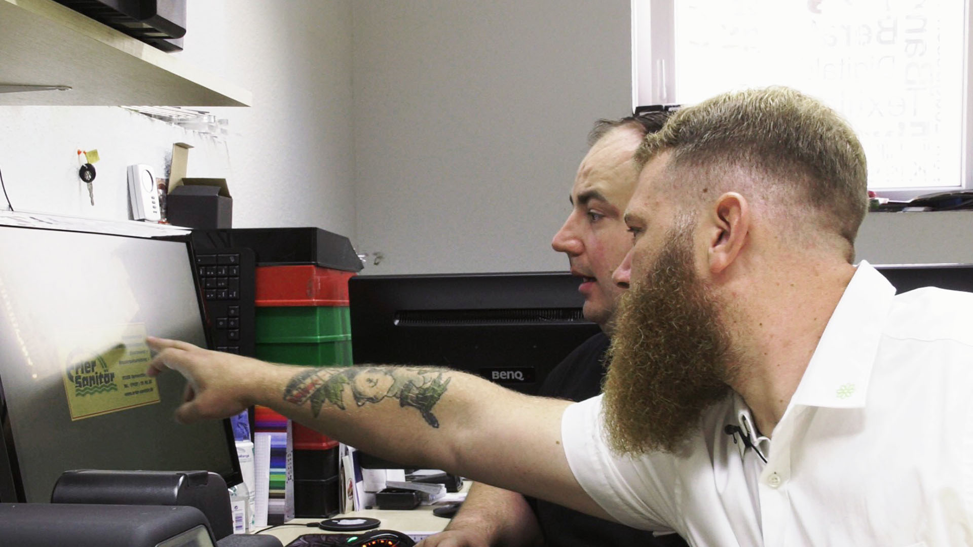 Zwei Männer mittleren Alters sitzen gemeinsam an einem Computer. Der eine weist mit der Hand auf den Bildschirm. Sein Unterarm ist stark tatowiert.