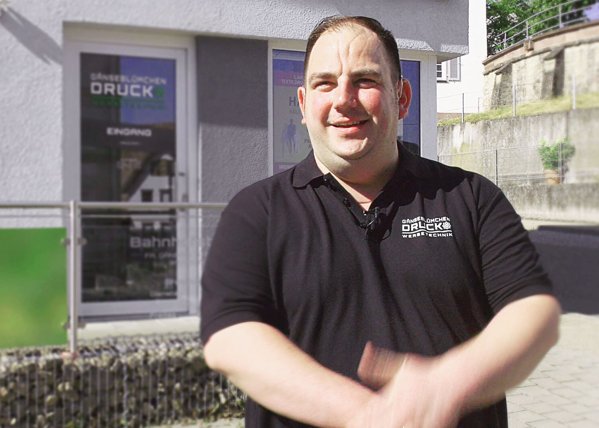 Ein untersetzter Mann mit schwarzem Polohemd mit weißer Applikation "Gänseblümchen Druck und Werbetechnik" steht vor der Druckerei.