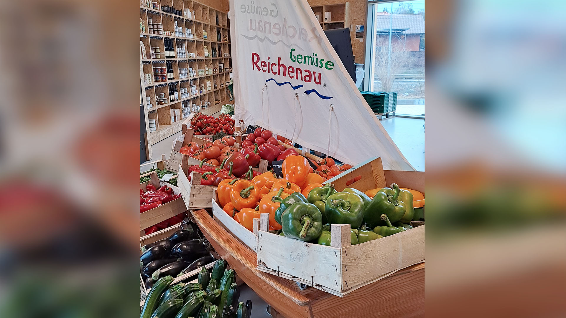 Ein kleiner Einhandsegler steht dicht gepackt mit Kisten voller reifer Tomaten, Paprika und Zucchini in der Markthalle.