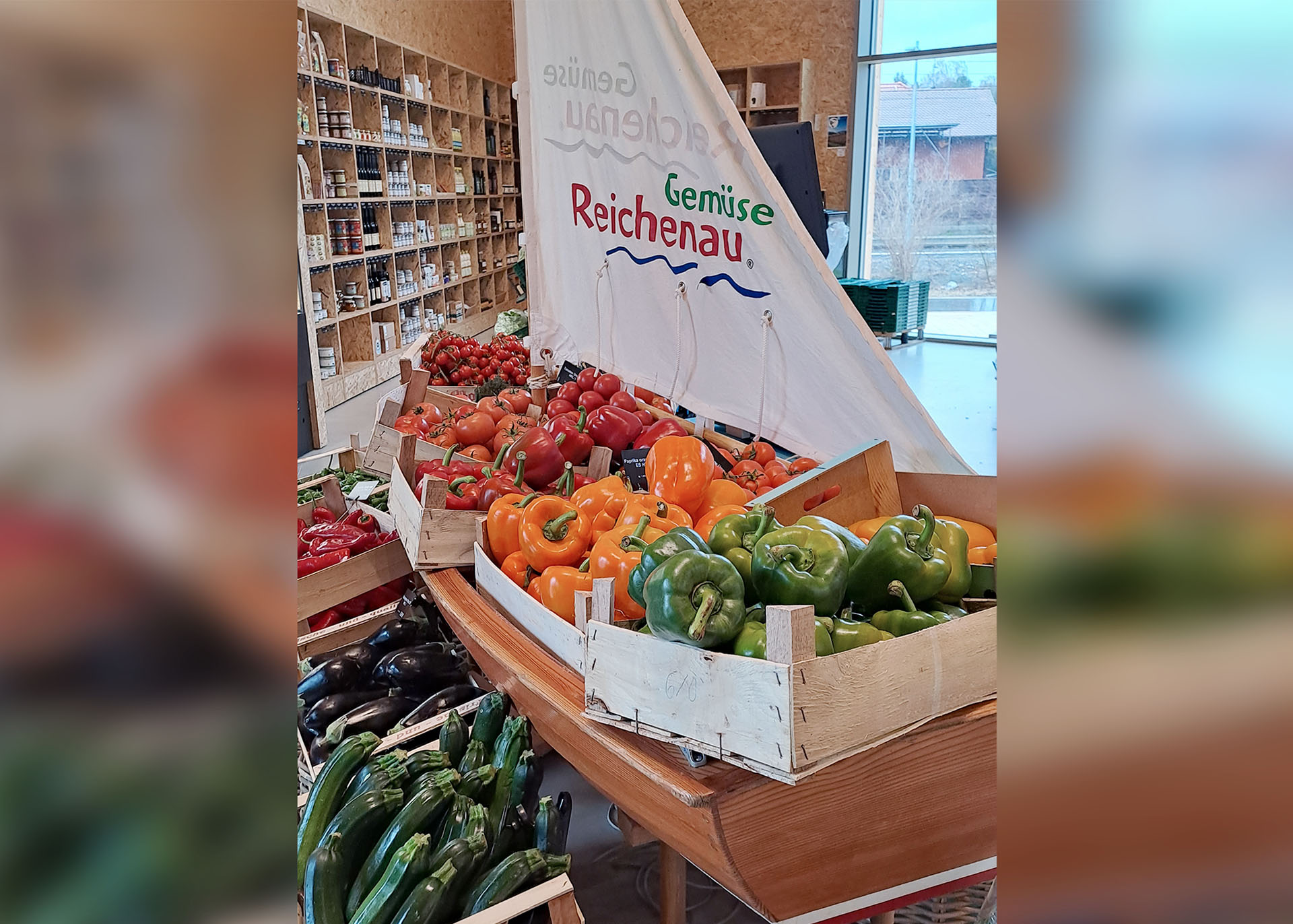 Ein kleiner Einhandsegler steht dicht gepackt mit Kisten voller reifer Tomaten, Paprika und Zucchini in der Markthalle.