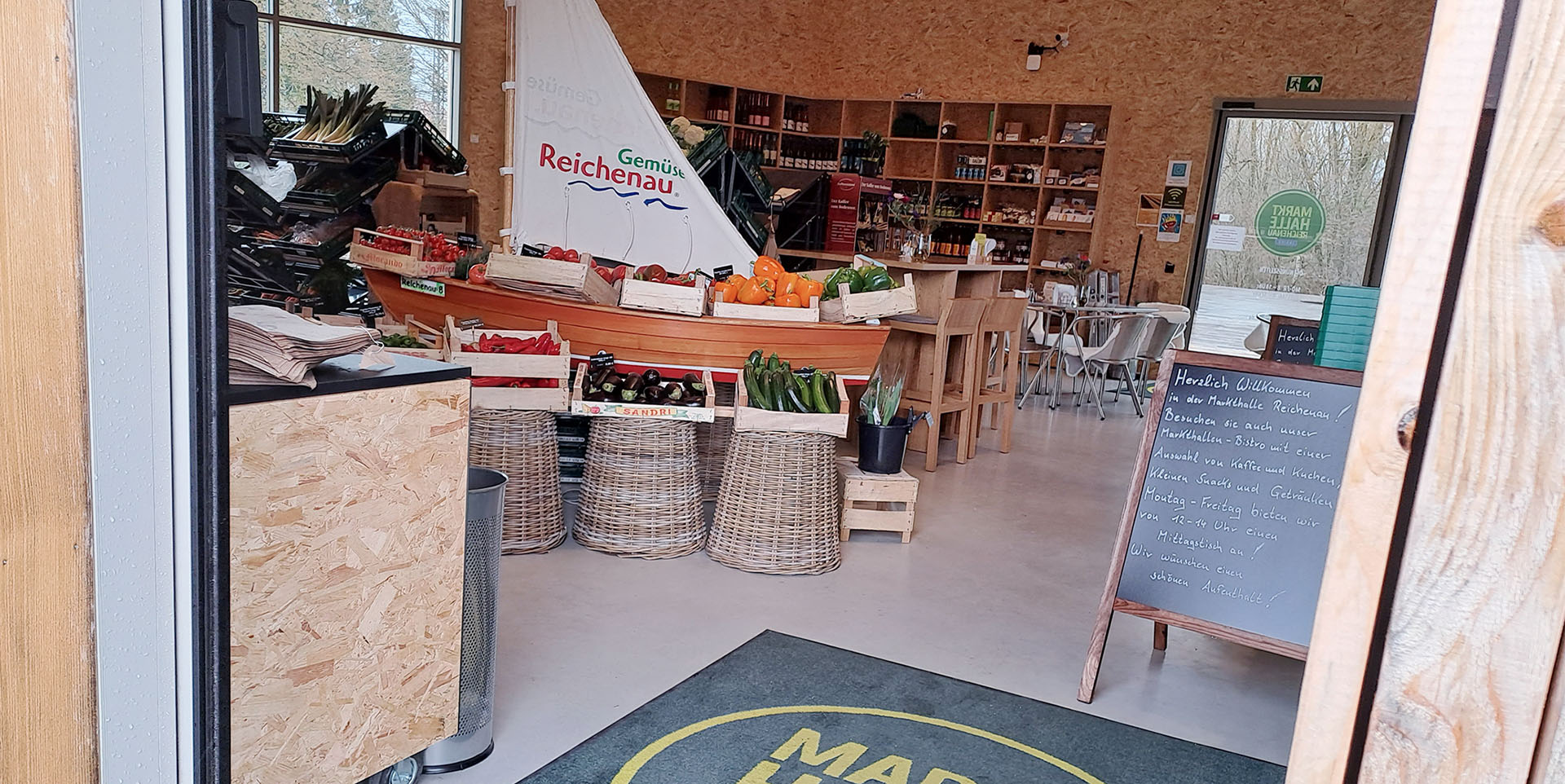Der Eingangsbereich der Markthalle Reichenau mit einem kleinen Segelboot voller Gemüsekisten. Auf dem Segel steht: "Gemüse Reichenau".