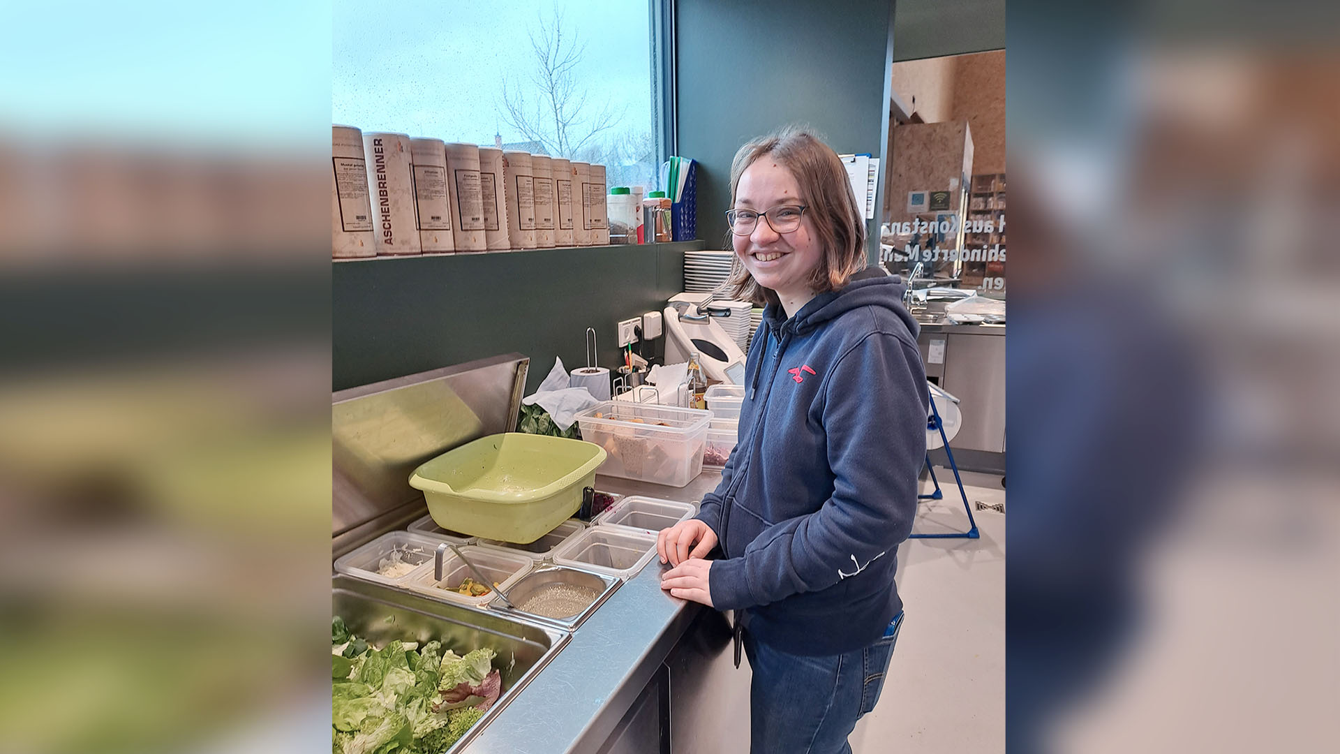 Eine junge Frau mit offenen schulterlangen Haaren und Brille steht an einer Theke mit vielen Schüsseln mit verschiedenen Zutaten für Salate.