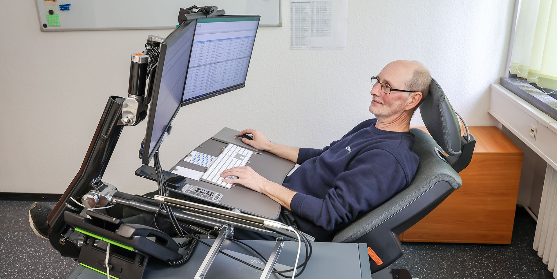 Schmerzfrei Arbeiten im Liegen Carsten Schönebeck an seiner Arbeitsstation mit zwei großen Monitoren, magnetischem Pult für Tastatur, Handy und Mousepad – hier in halb liegender Position.