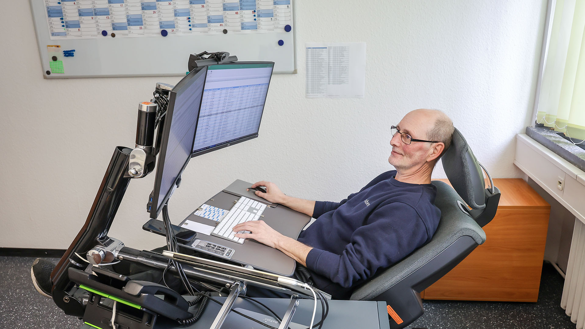 Carsten Schönebeck an seiner Arbeitsstation mit zwei großen Monitoren, magnetischem Pult für Tastatur, Handy und Mousepad – hier in halb liegender Position.