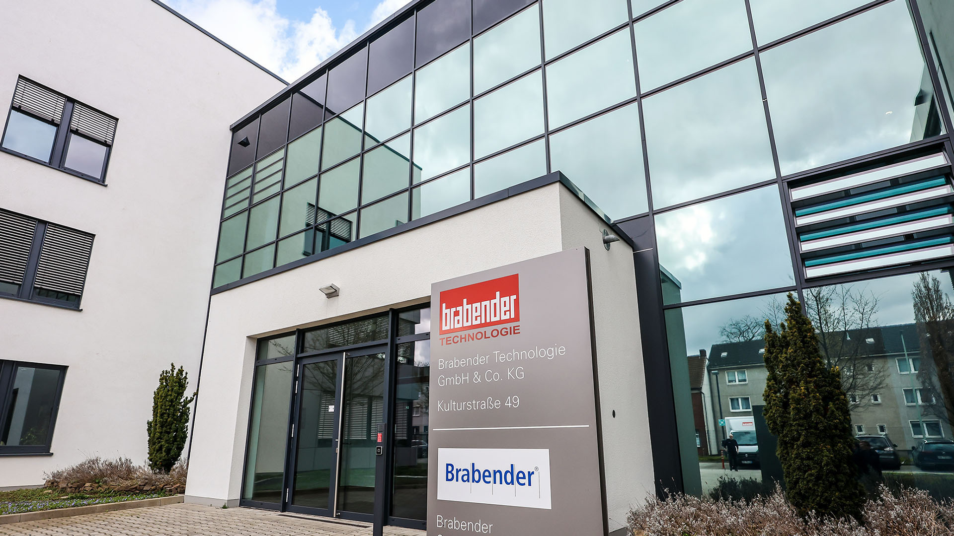 Foto vom Eingang des Hauptfirmensitzes der Brabender GmbH & Co. KG. Es ist ein modernes, aus zwei großen würfelartigen Blöcken zusammengesetztes Gebäude. Der rechte Würfel hat eine hohe Glasfront.