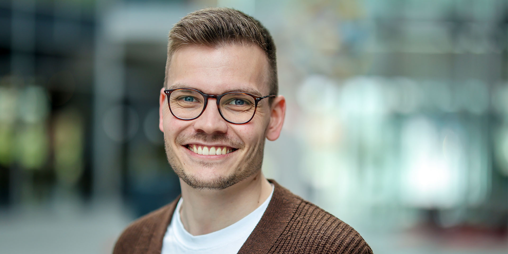 Jonas Potthoff, LVR-Inklusionsamt Porträt von Jonas Potthoff, ein junger Mann mit kurzem modernen Haarschnitt, Hornbrille, Drei-Tage-Bart und einem strahlenden Lächeln. Im unscharfen Hintergrund ist die Glasfront eines Bürogebäudes zu sehen.