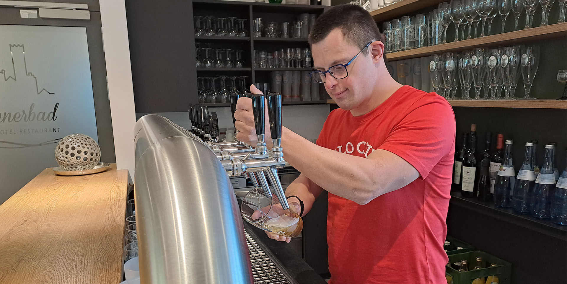 Lukas Geser steht im roten T-Shirt am Zapfhahn der Hotelbar und zapft mit konzentriertem Blick ein Bier.