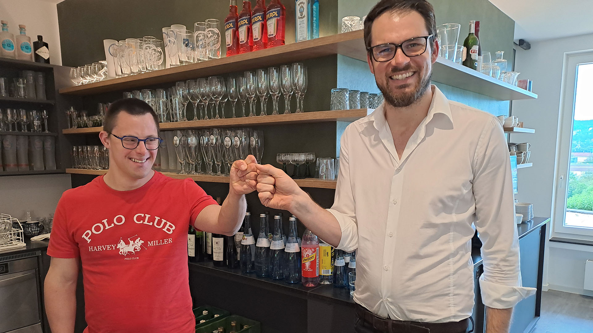 Hotelleiter Dominik Buck im weißen Oberhemd und Lukas Geser (links) im roten T-Shirt stehen mit aneinander gelegten Fäusten an der Hotelbar..